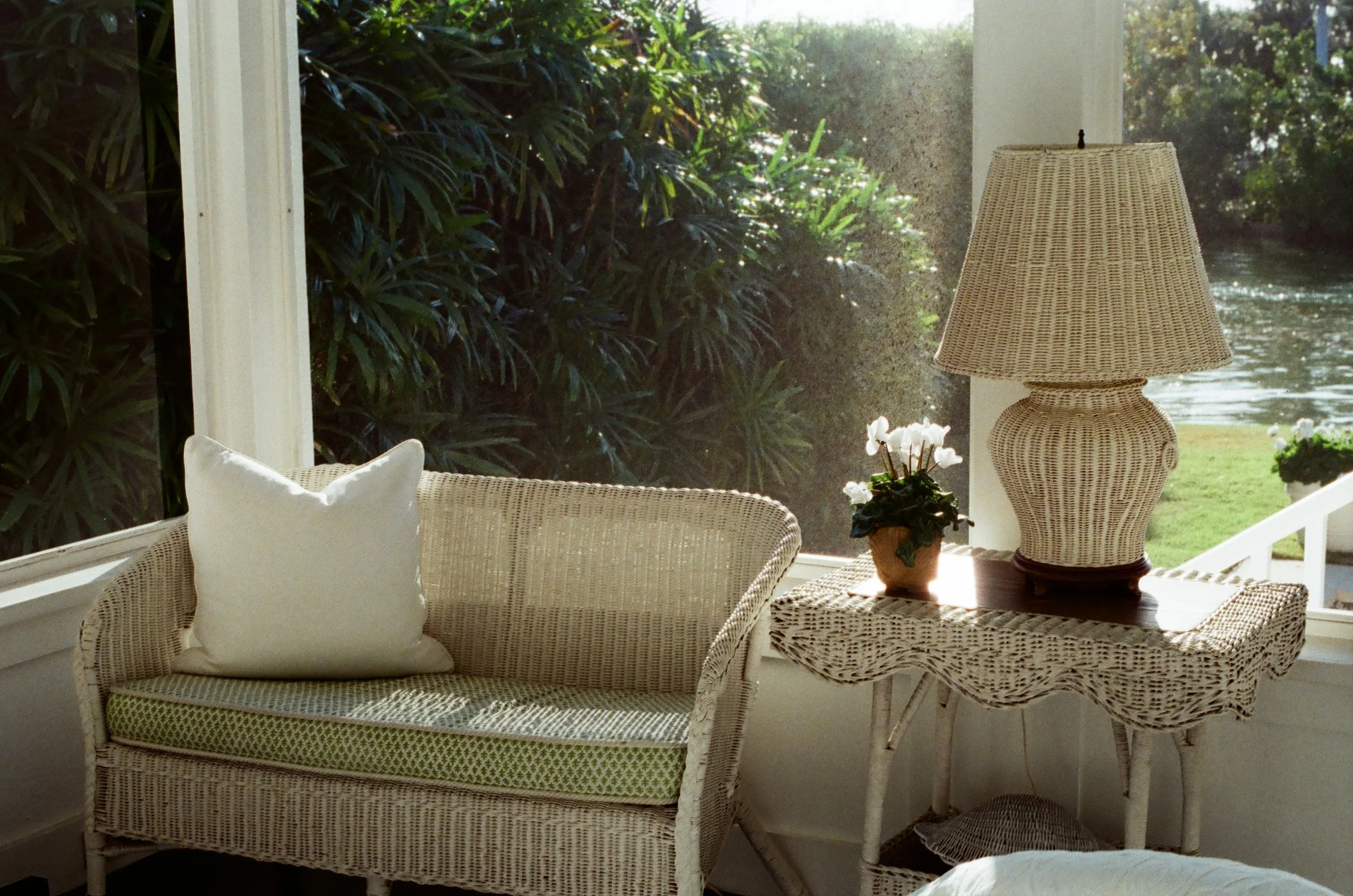 Sunlit interior of a cozy room with a wicker loveseat that has a white pillow and green cushion, next to a wicker table holding a large wicker lamp and a potted white flower. Large windows reveal lush green foliage and a body of water outside.