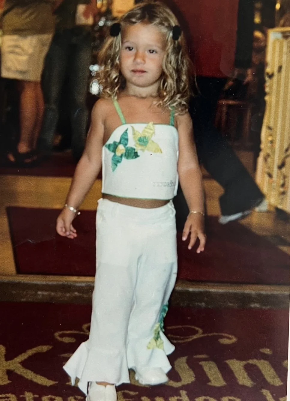 A young girl with curly blonde hair standing indoors, wearing a white two-piece outfit with green leaf designs and sandals, with a carpeted floor and blurred people in the background.