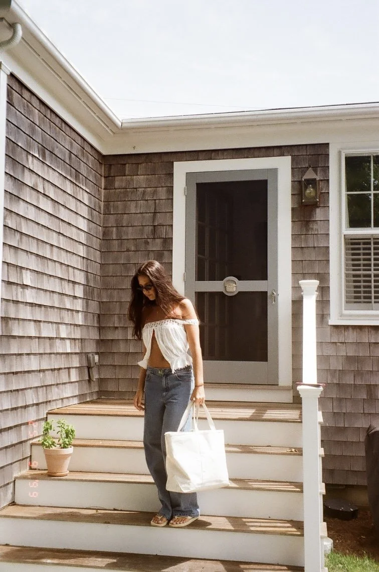 Young woman with long brown hair wearing sunglasses, a white off-the-shoulder top, and blue jeans, standing on wooden stairs outside a house, holding a large white tote bag.