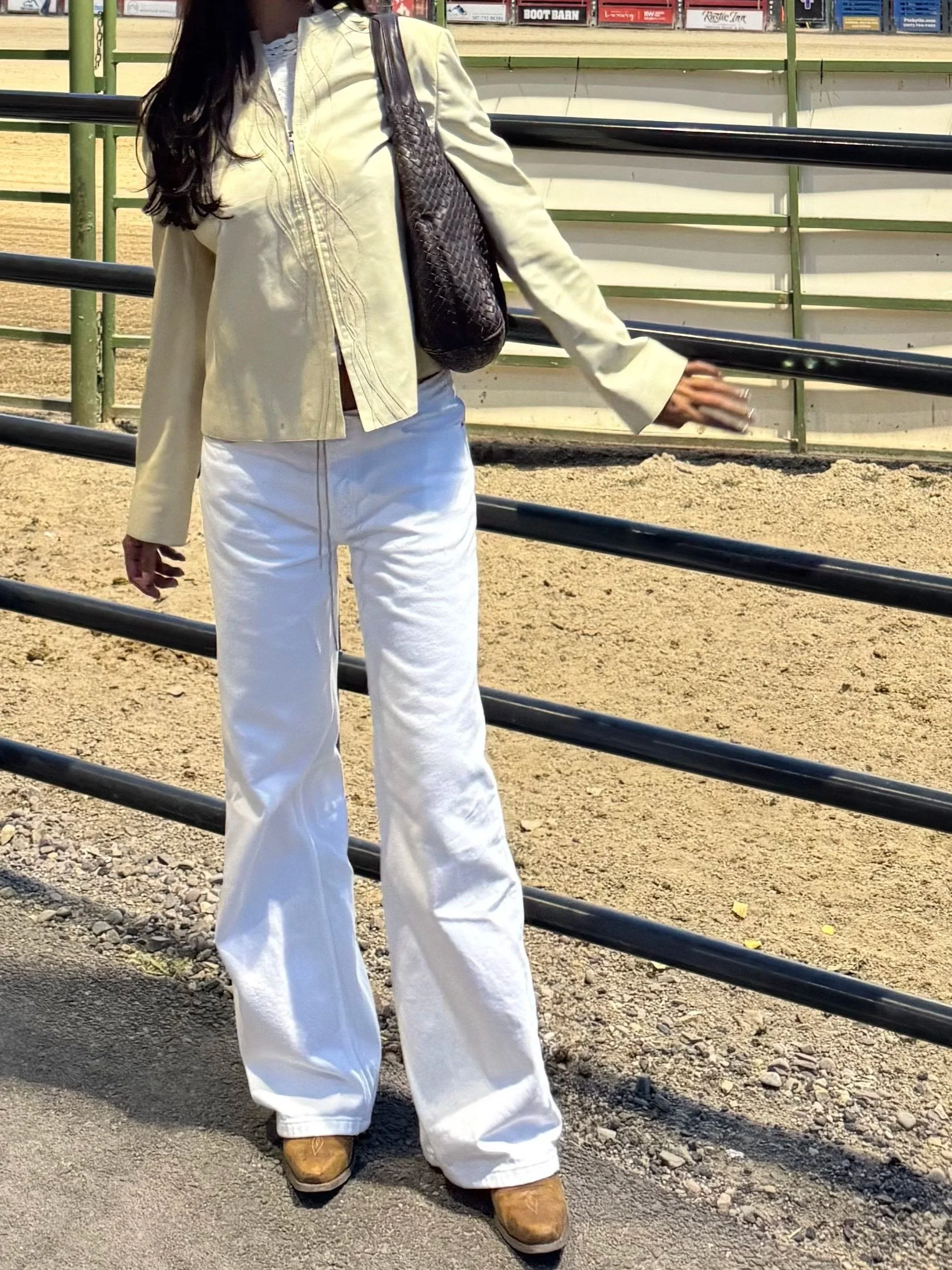 A woman standing outdoors near a fenced area, wearing a light-colored jacket, white wide-leg pants, tan boots, and carrying a dark textured shoulder bag.