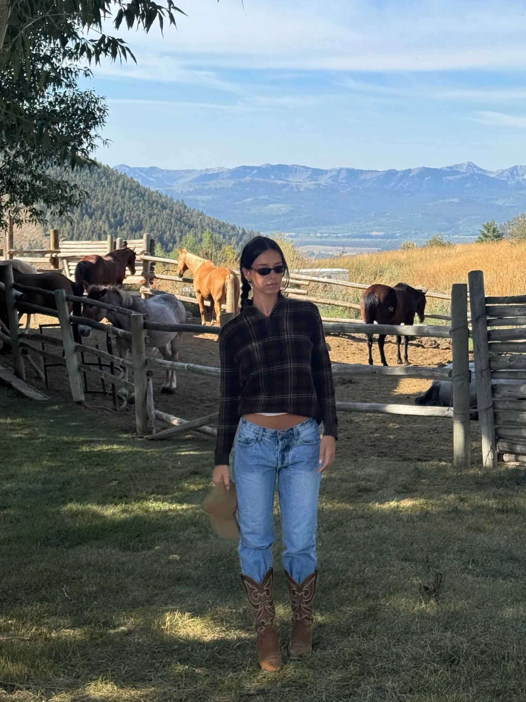 A woman with dark hair, wearing sunglasses, a plaid shirt, jeans, and cowboy boots, standing on grass near a wooden fence with horses in the background, mountains and blue sky in the distance.