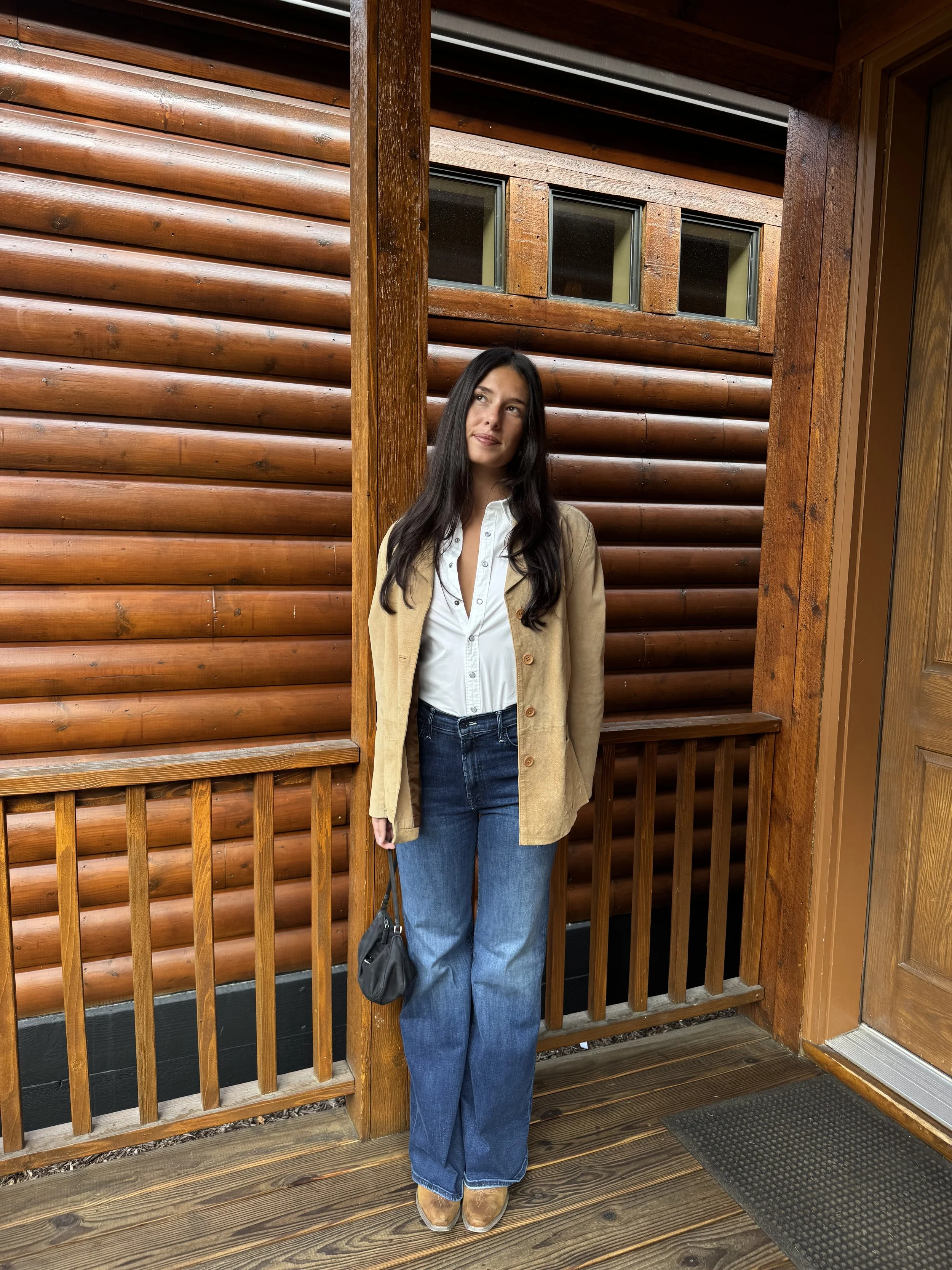 A woman with long dark hair wearing a beige coat, white button-up shirt, and blue jeans, is standing on a wooden porch with a log cabin-style wall behind her.