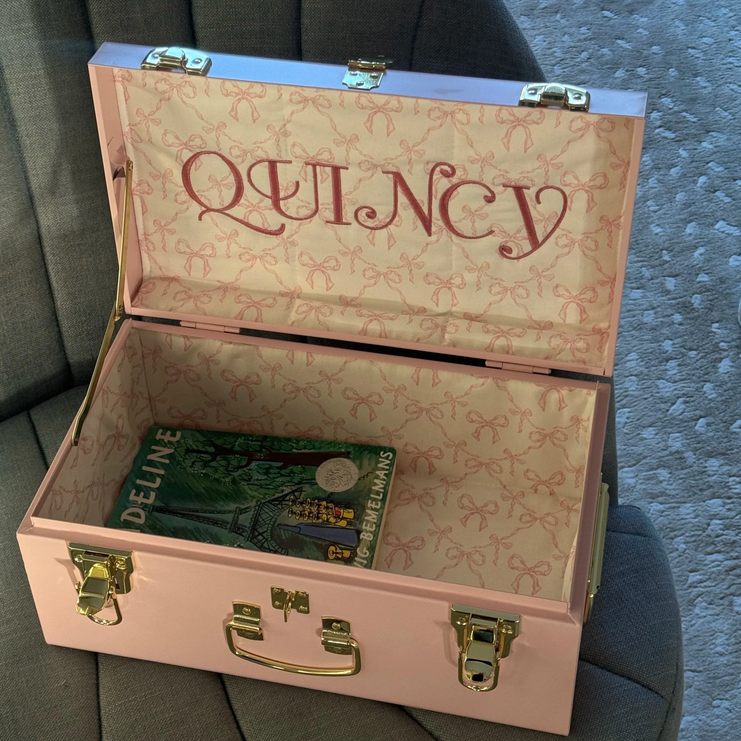 Open pink jewelry box with gold clasps and handle, interior lined with pink ribbon bow pattern, containing a vintage green book titled 'WONDERFUL BEMEMANS', resting on a gray fabric surface.