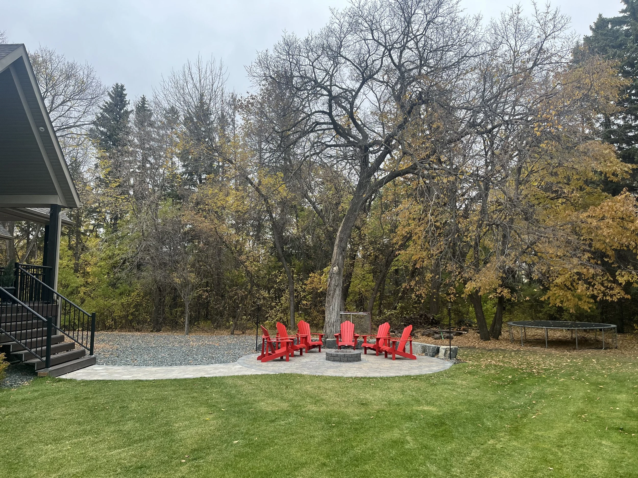 An outdoor backyard scene with red Adirondack chairs arranged in a circle around a tree trunk on a stone patio. There is a house on the left side and a trampoline on the right side, with trees in fall foliage in the background.