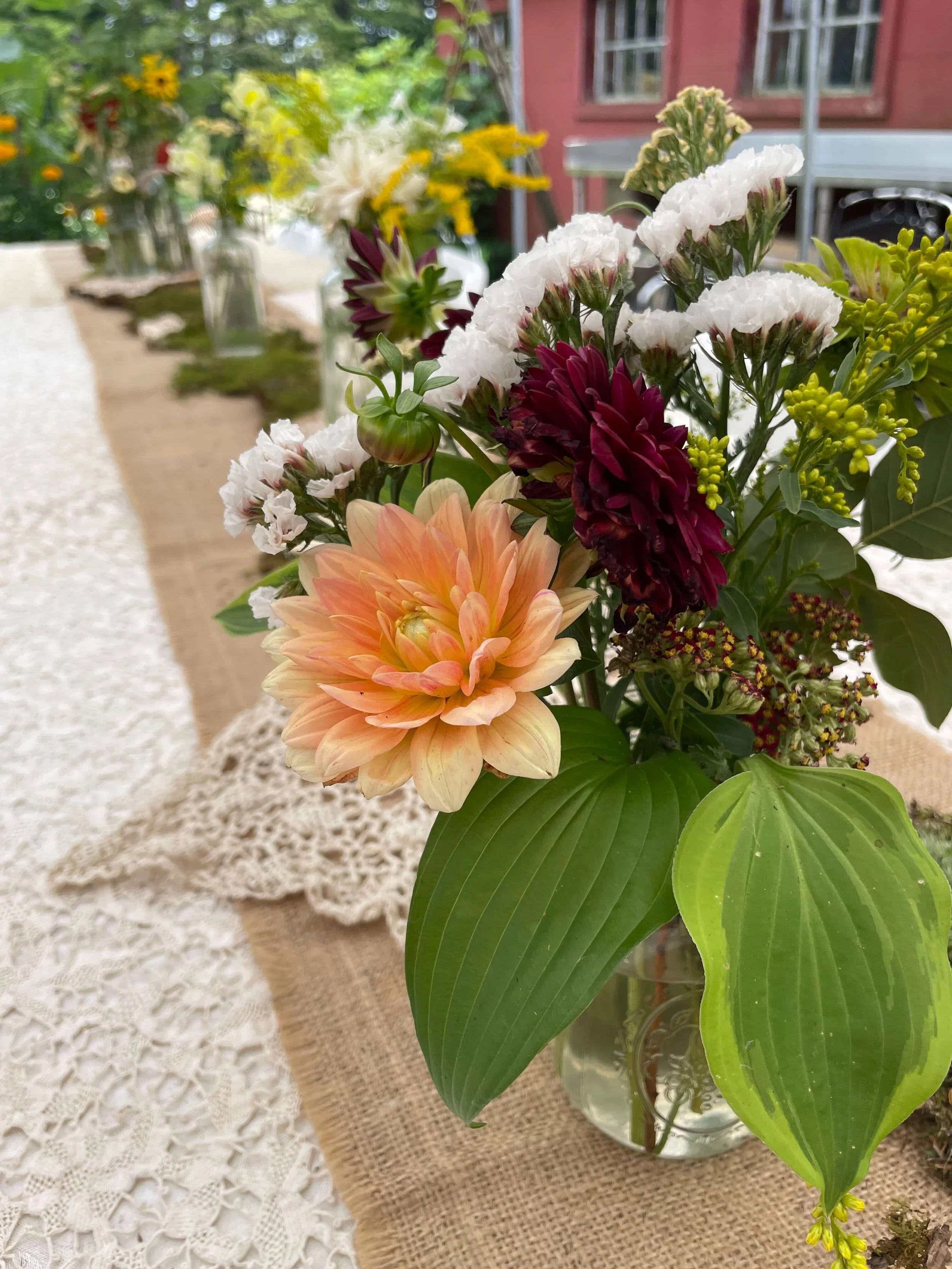 Close-up of a flower arrangement with a peach dahlia, white, maroon, and white flowers, yellow filler flowers, and large green leaves on a table with lace and burlap tablecloths, outdoors with flowers and a building in the background.