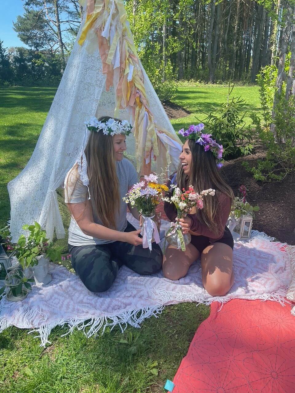 Two women wearing flower crowns sit on a blanket outdoors, exchanging flowers and smiling. They are surrounded by greenery and sunlight, with a decorative lace tent behind them.
