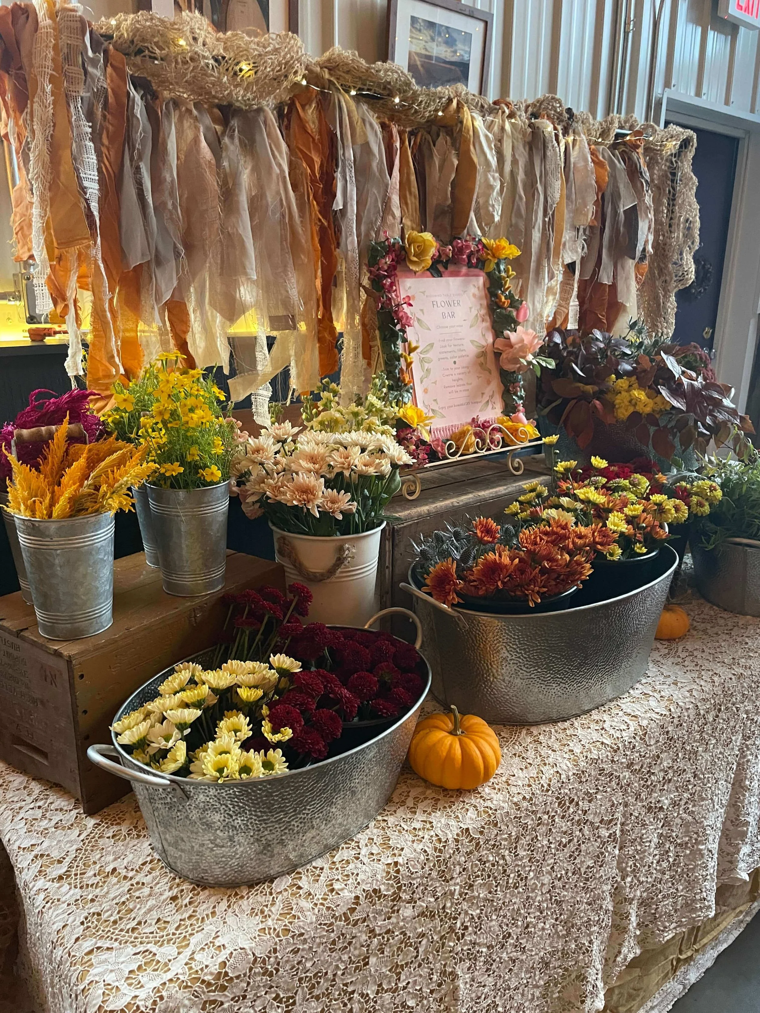 Display of various potted and arranged autumn flowers, including chrysanthemums and daisies, on a lace-covered table along with small pumpkins. Behind the flowers is a decorative backdrop made of ribbons and fabric pieces, and there is a framed menu 