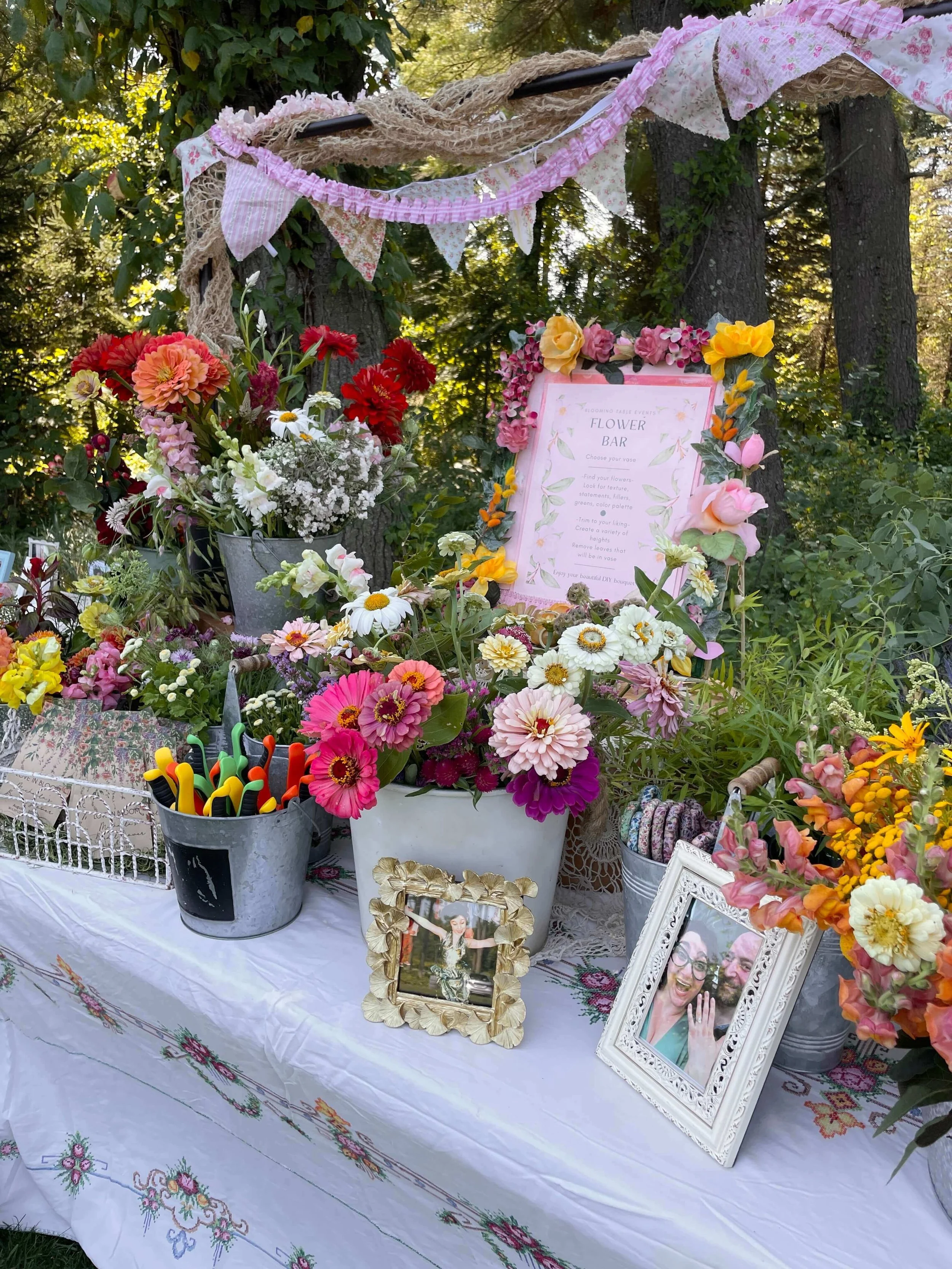 A garden table decorated with colorful flowers, framed photographs, and a sign that says 'Flower Bar' amidst trees and greenery.