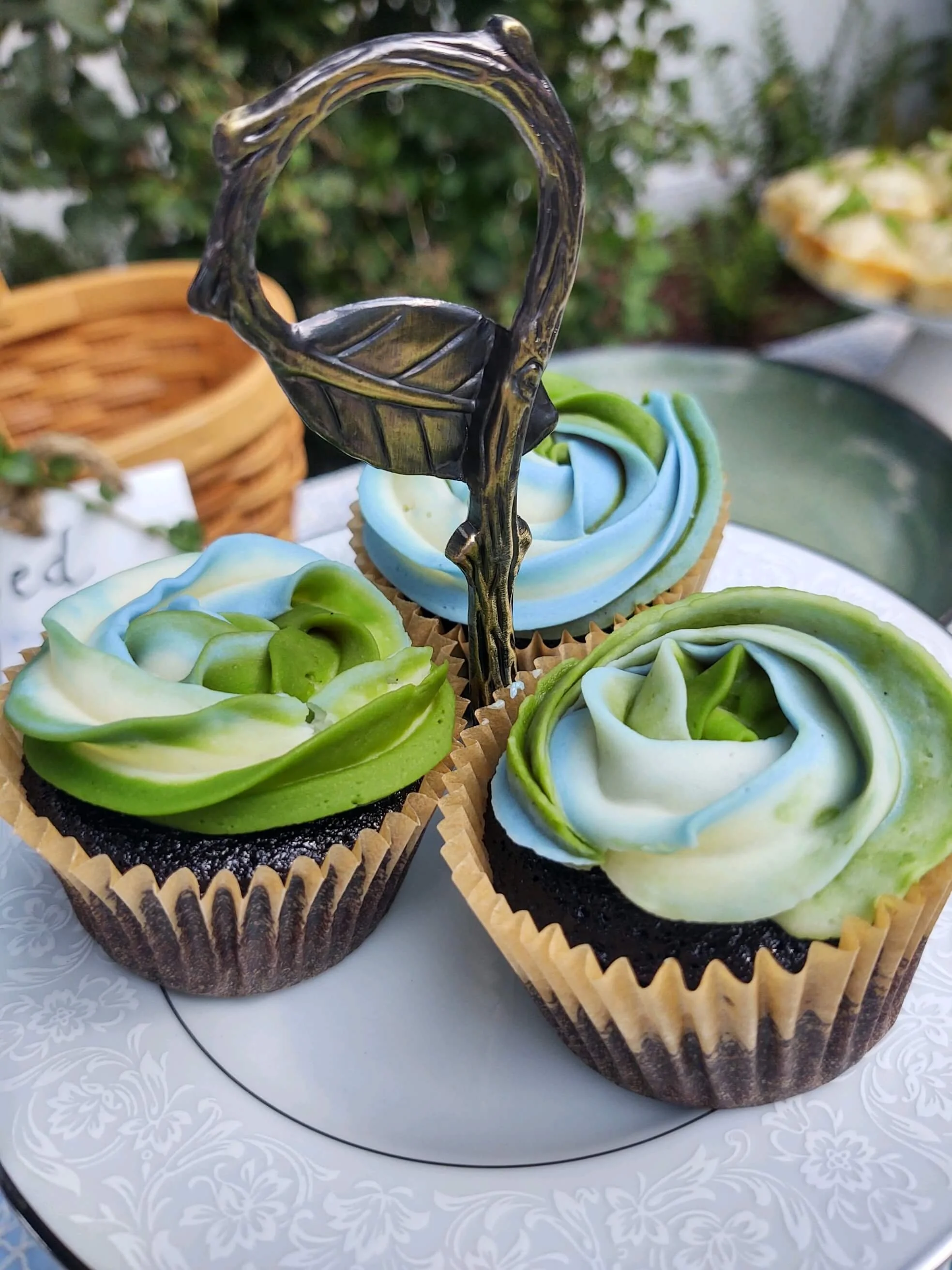 Three cupcakes with blue, green, and white swirl frosting on a decorative white plate outdoors.
