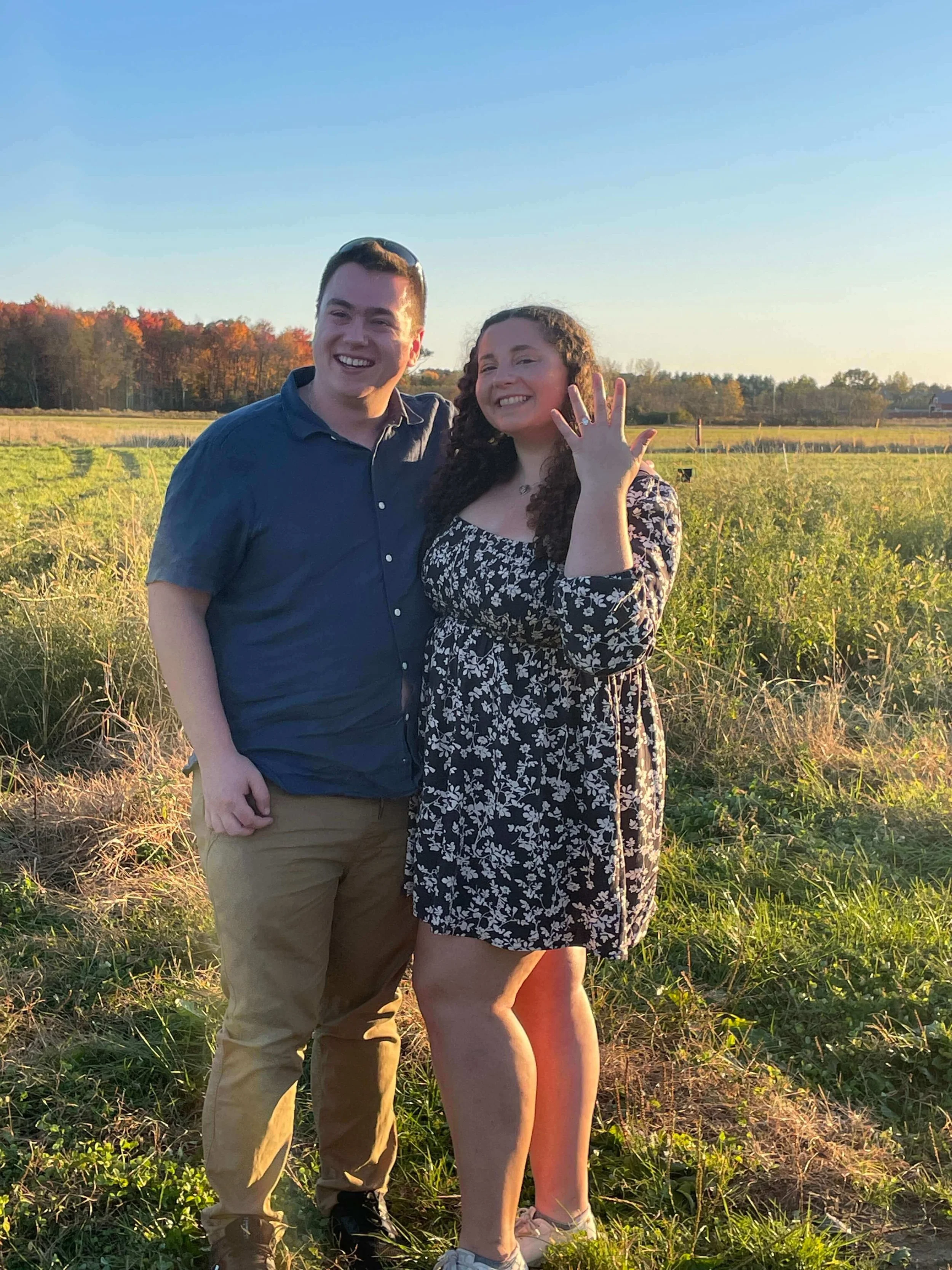 A young man and woman standing outdoors in a field, smiling at sunset. The woman is showing off a ring on her left hand, indicating she is recently engaged.