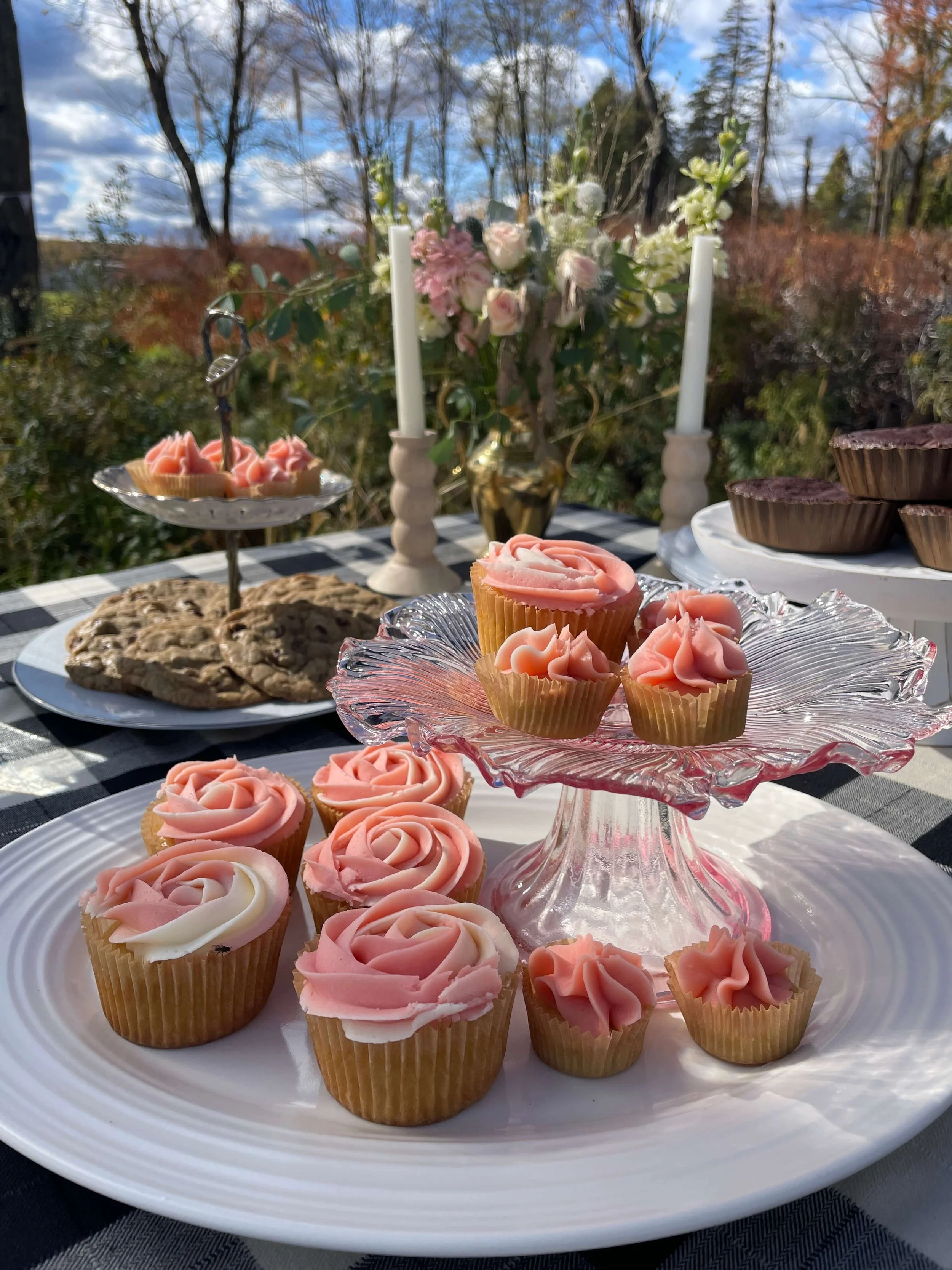 Pink and white frosted cupcakes on a glass cake stand and white plate, with cookies, in an outdoor setting with trees, flowers, candles, and a cloudy sky in the background.