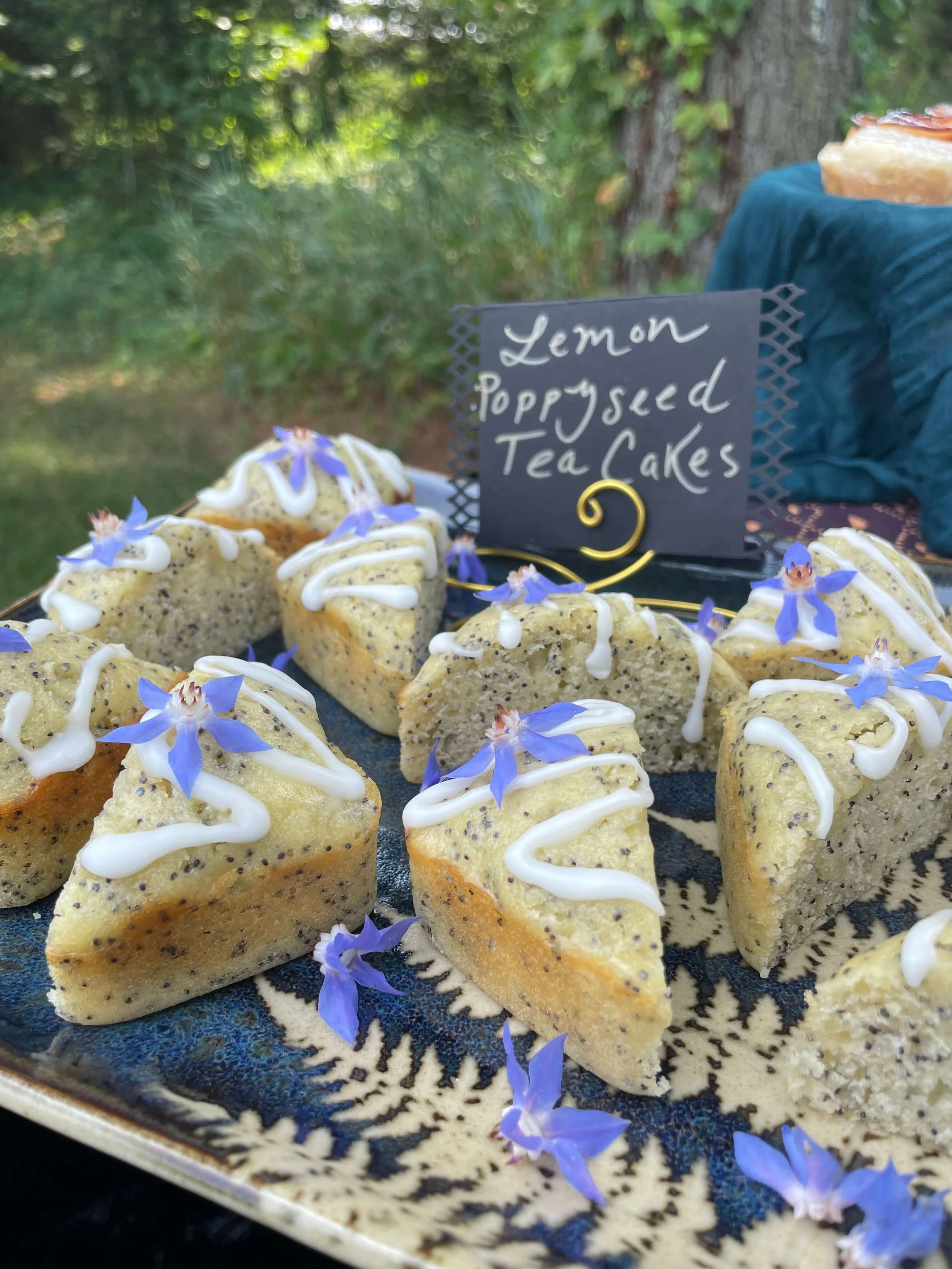Lemon poppy seed tea cakes decorated with white icing and blue flowers, displayed on a decorative plate outdoors.