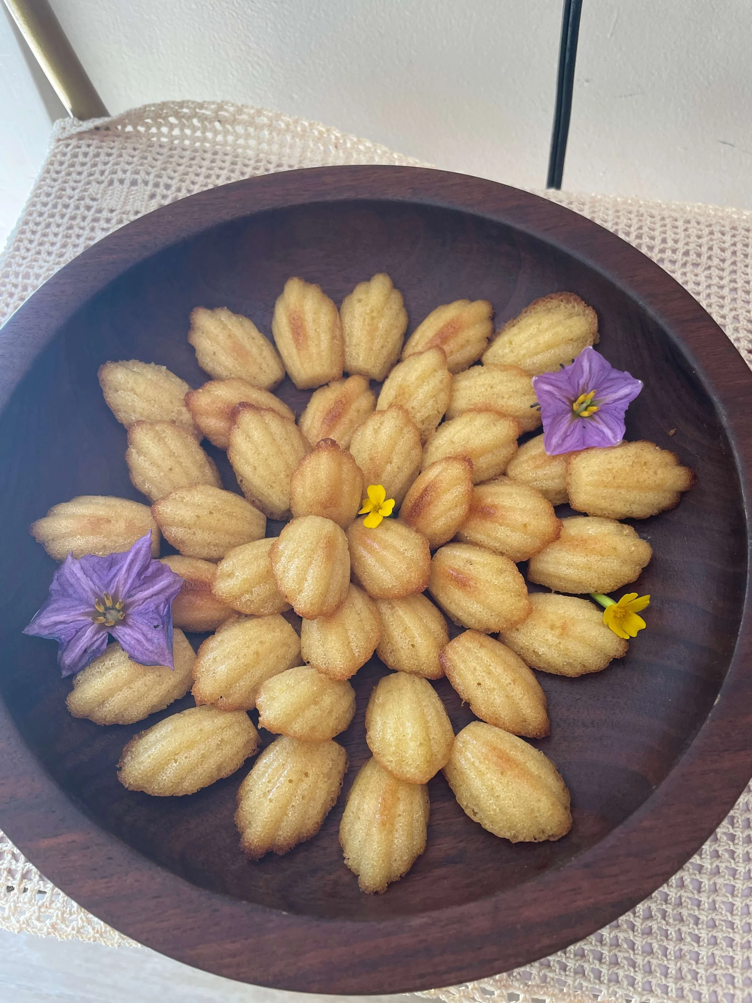 A wooden bowl filled with small Madeleine cookies, garnished with purple and yellow flowers.