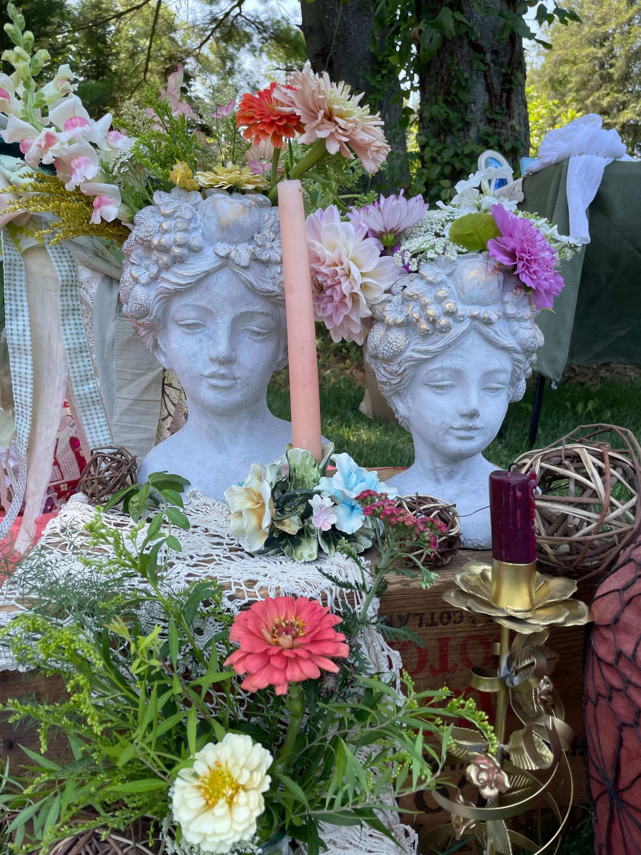 Two white bust statues with floral arrangements on their heads, surrounded by colorful flowers, candles, and decorative items on a table at an outdoor setting.