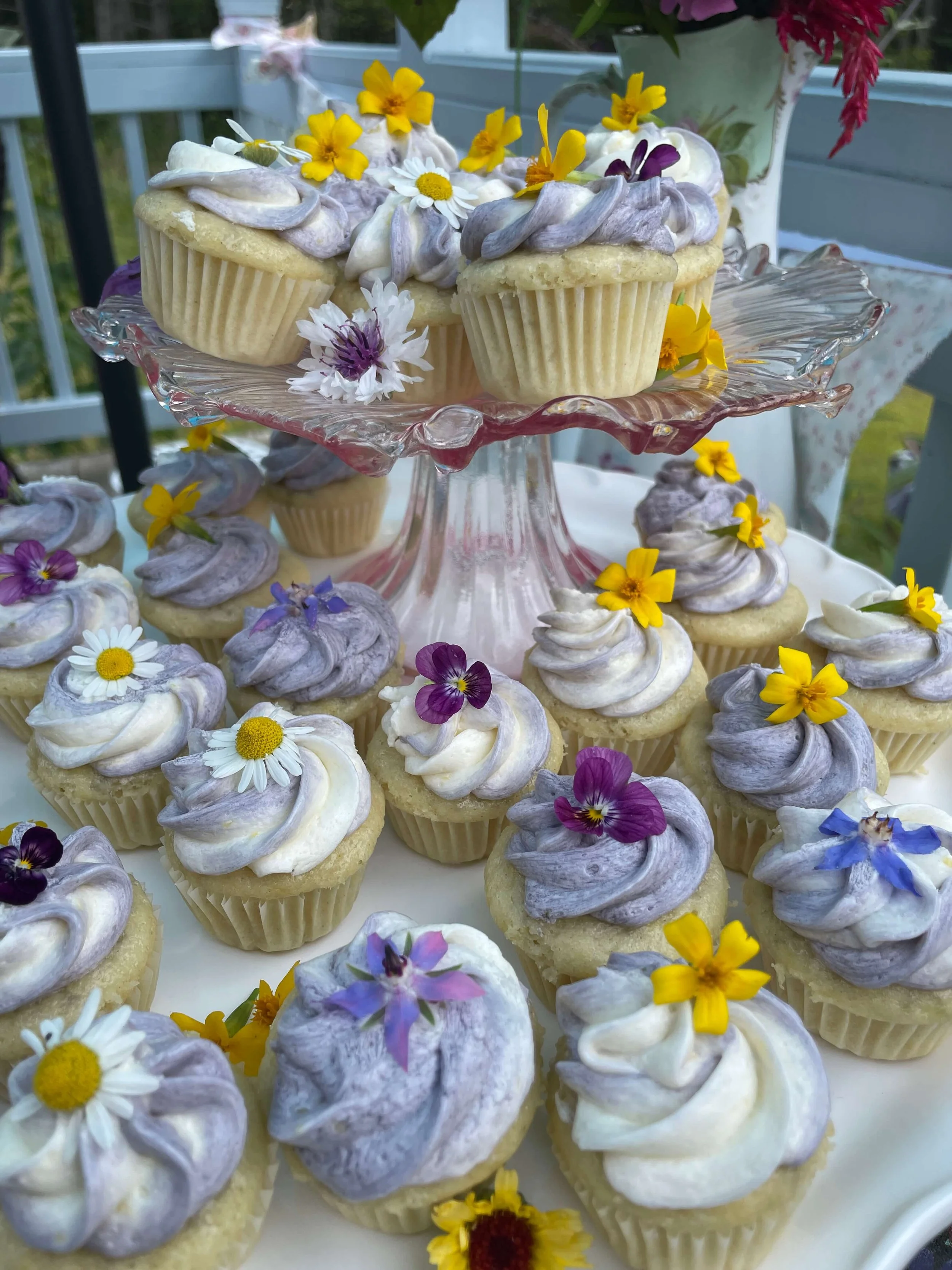 A display of cupcakes topped with purple and white swirled frosting, decorated with edible flowers in yellow, purple, and white on a glass tiered stand and surrounding tray.