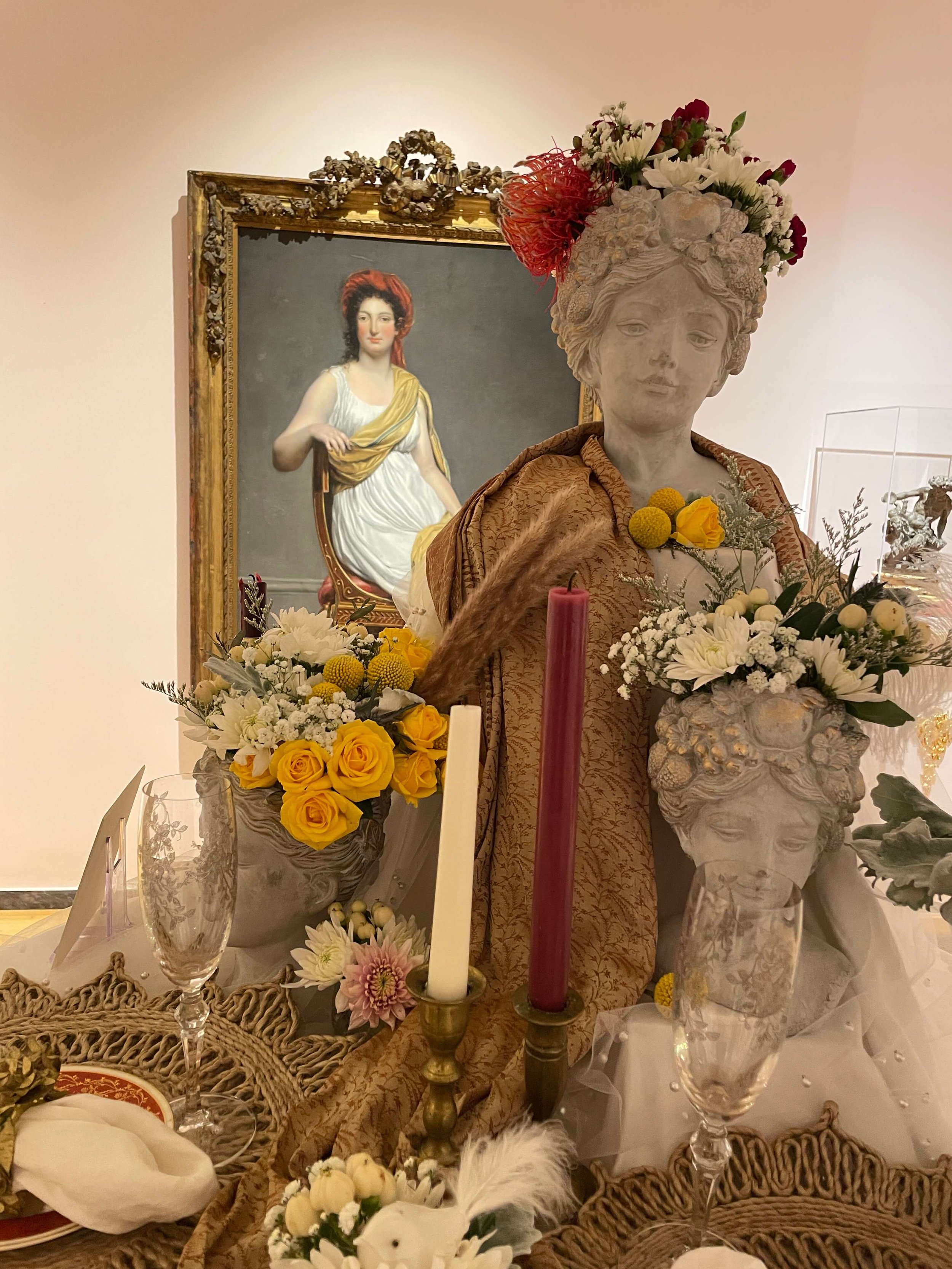 A decorated display featuring a bust sculpture with flowers and candles, and a painting of a woman with red hair in the background.