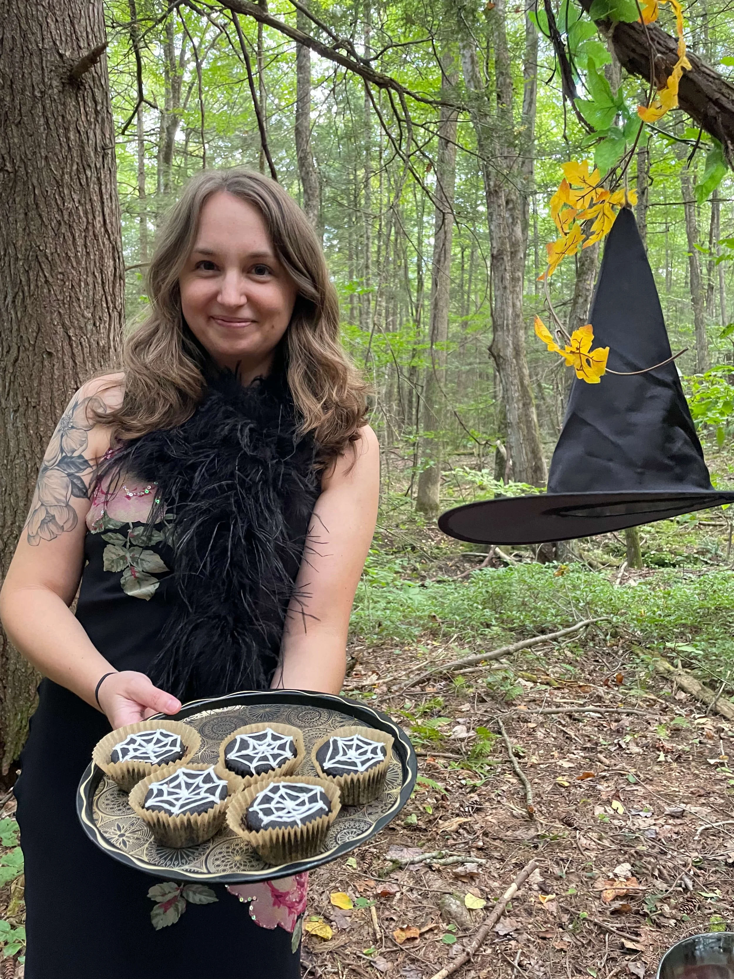 A woman in a black dress with floral details, holding a tray of cupcakes decorated with spiderweb icing, standing in a forest next to a tree. A witch’s hat is hanging from a branch nearby.