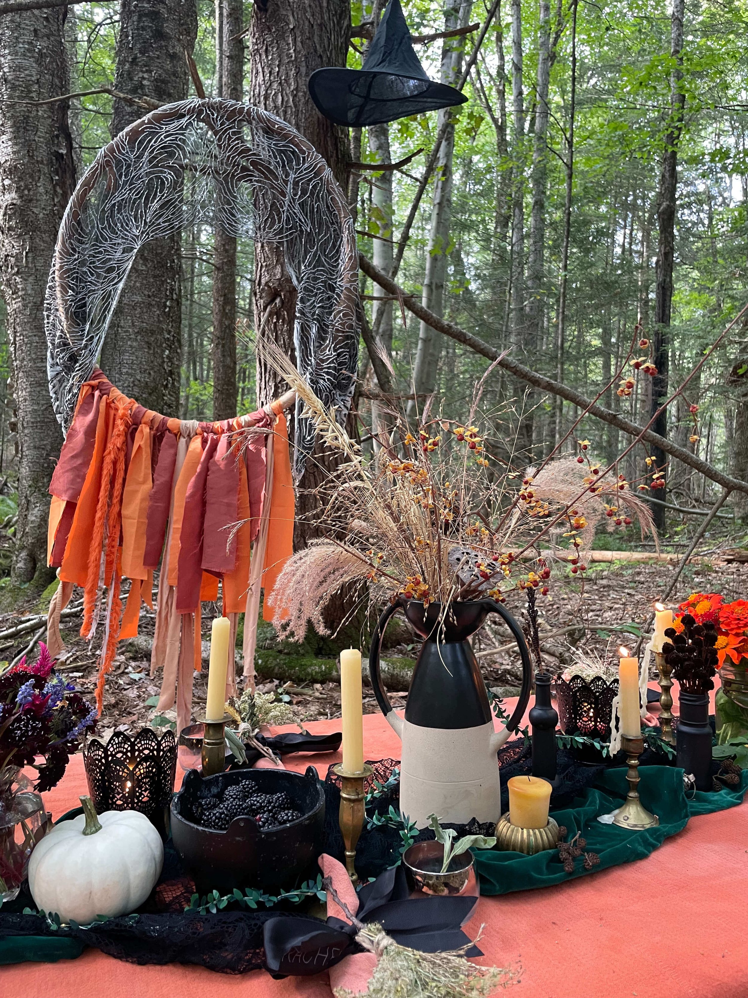 An outdoor picnic set up in a forest with candles, a black bowl filled with blackberries, and various flowers and decorations, including a white pumpkin, a black hat, a veil, and a ribbon with fall colors. Trees and greenery surround the scene.