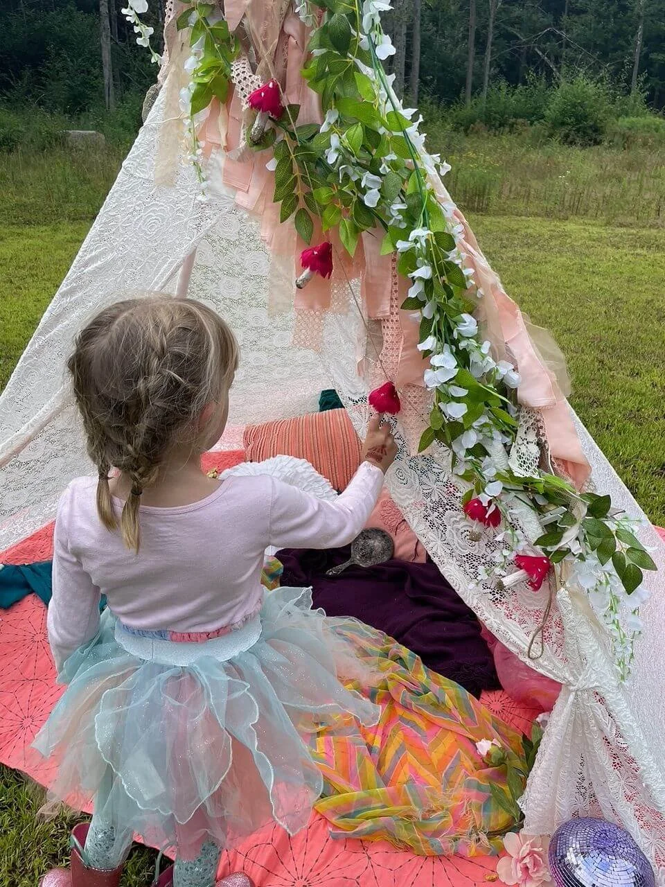 A young girl with braided hair and a colorful tutu is inside a small, white lace tent decorated with flowers and greenery. She is reaching out to touch a red flower hanging from the tent.