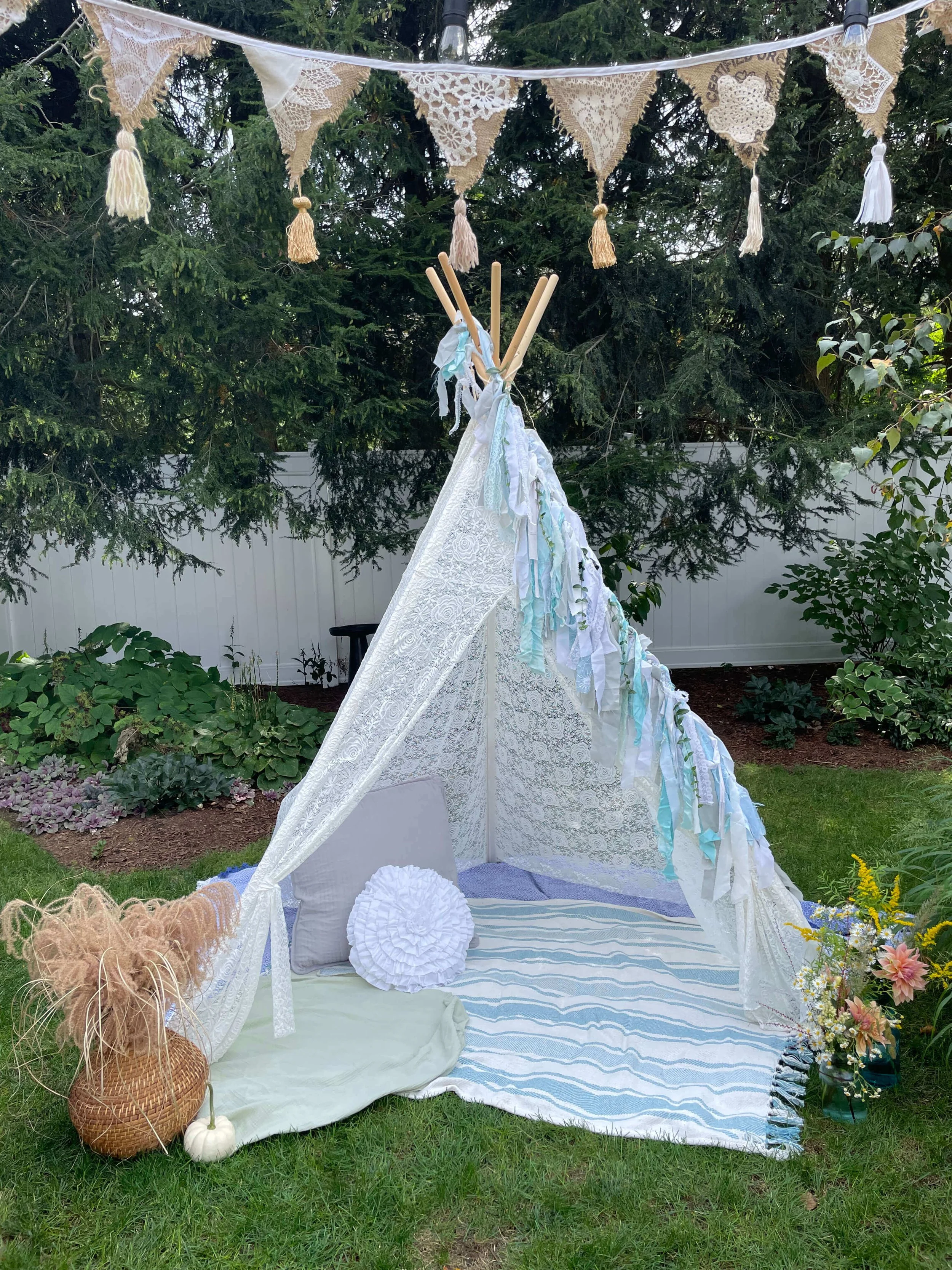 A backyard scene with a small white lace tent decorated with pastel-colored ribbons. Inside the tent, there are cushions and blankets, with a vase of flowers nearby. Above, a string of lace and tassel banners is hanging, and a bundle of pampas grass 