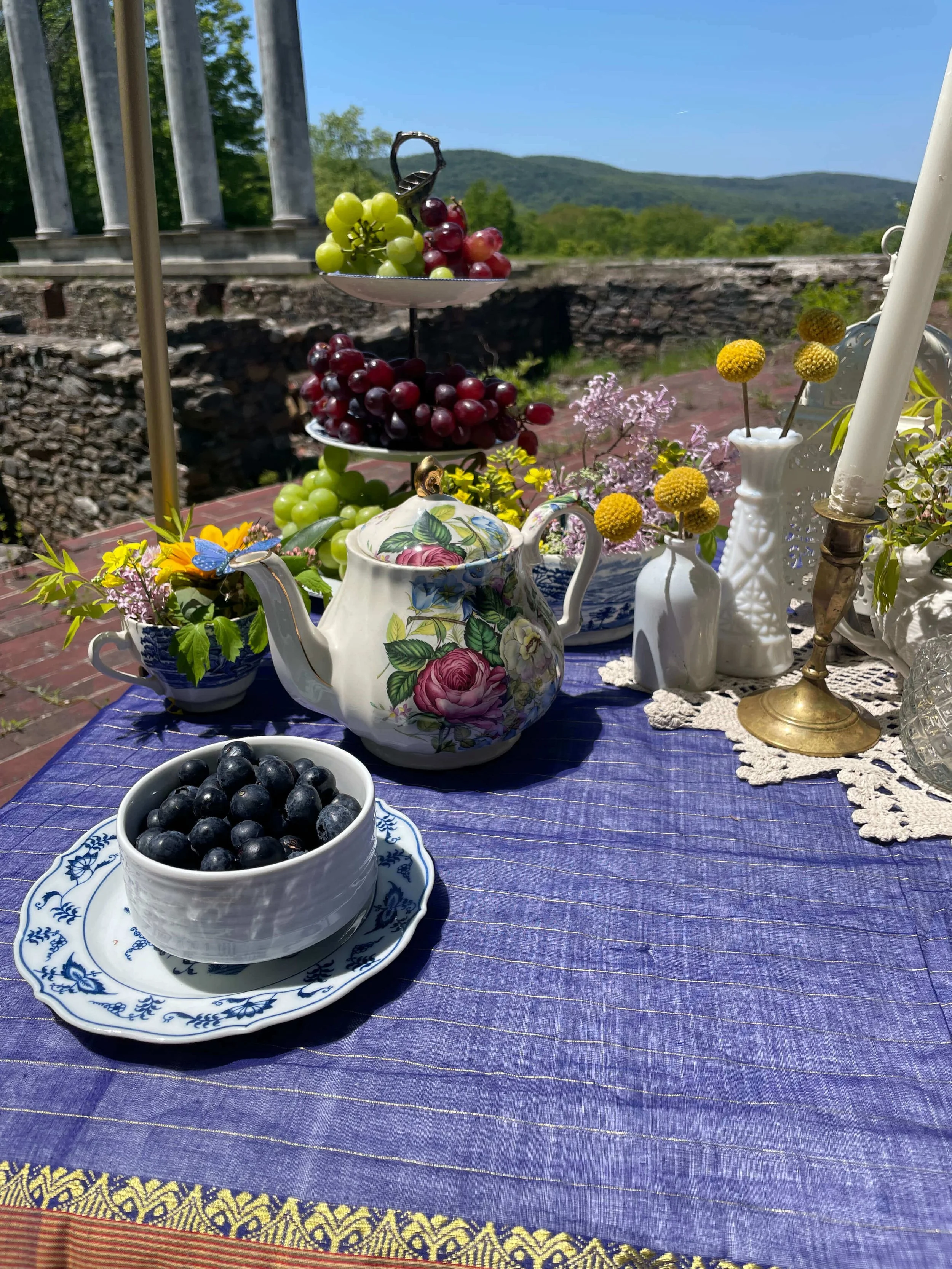 A table outdoors set with a purple tablecloth, a floral teapot, small vases with yellow and purple flowers, bowls with green grapes, and a tiered stand with green and red grapes. Background shows green trees, stone wall, and distant hills under a blu