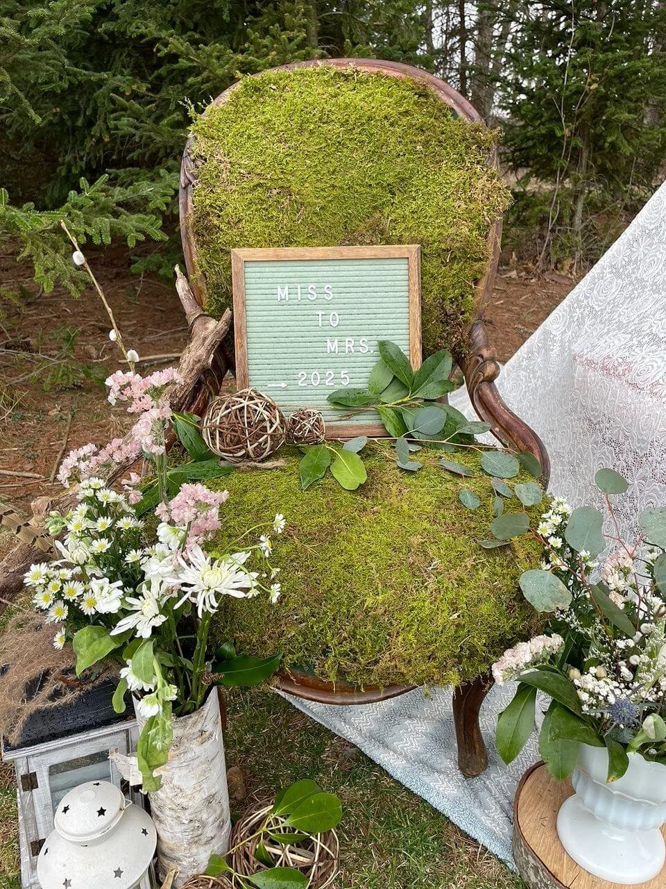 A moss-covered vintage chair with wooden arms, placed outdoors amid greenery, decorated with pink and white flowers, leafy branches, and decorative wicker balls. A small sign reads 'Miss To Mrs. 2025'.