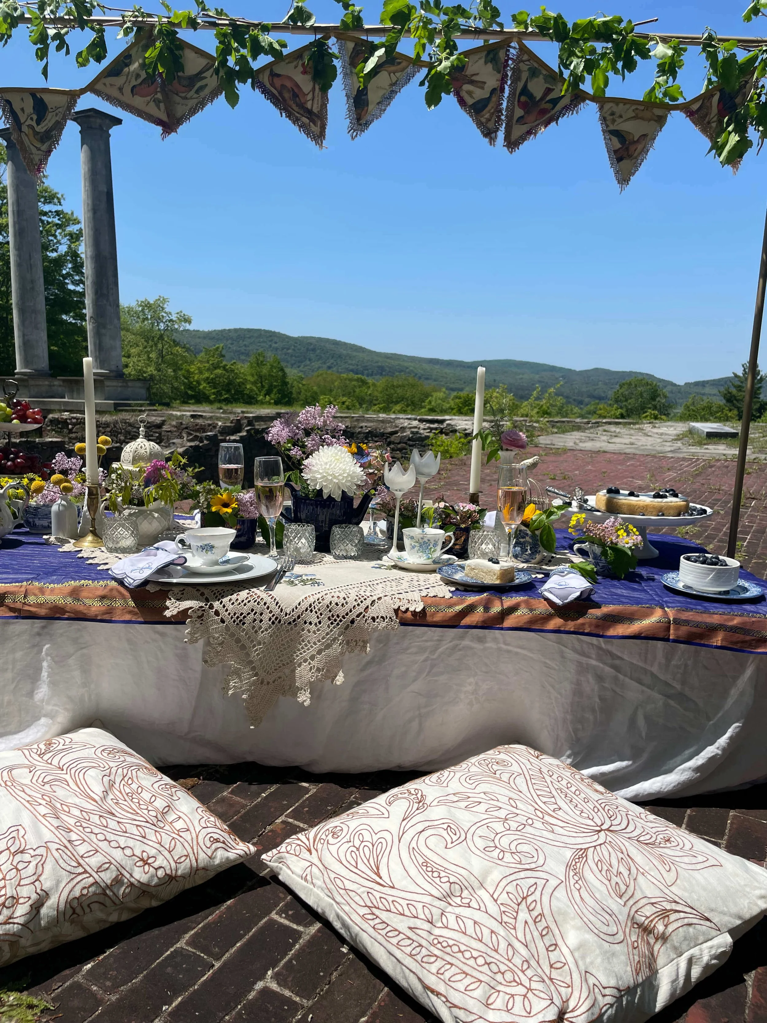 Outdoor picnic table set with floral decorations, tableware, and food, under a canopy with scenic mountain views in the background.