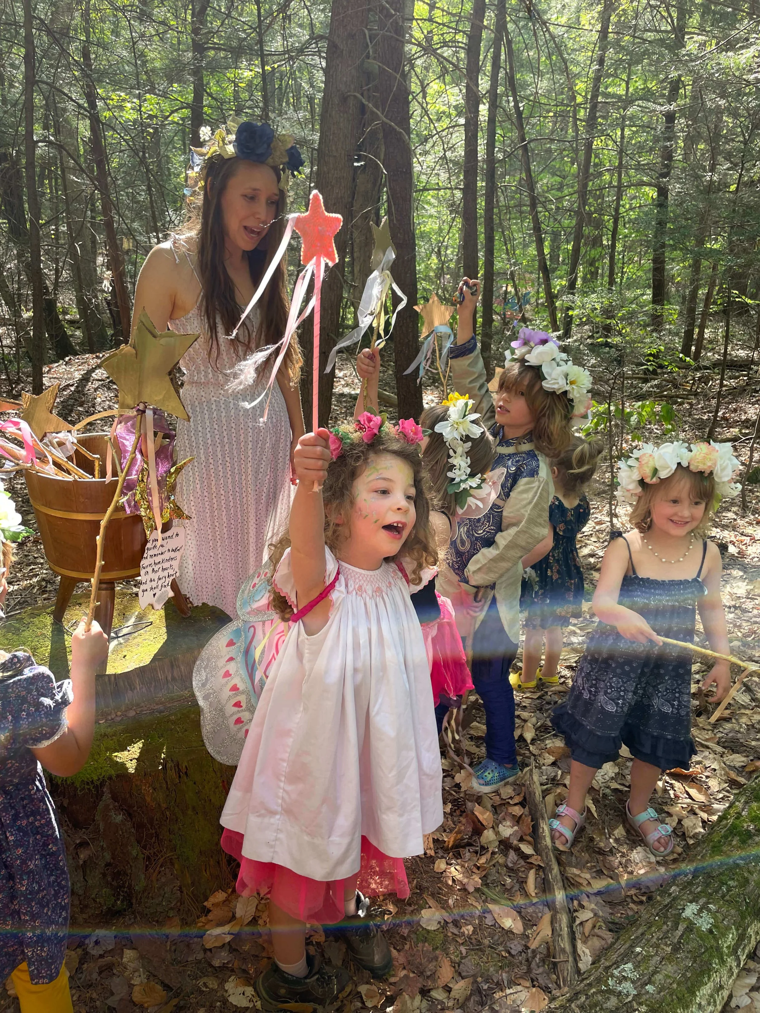 A woman dressed as a fairy with a flower crown and fairy wings, surrounded by children in a forest during a fairy-themed event. The children are dressed in fairy costumes with flower crowns and are holding wands.