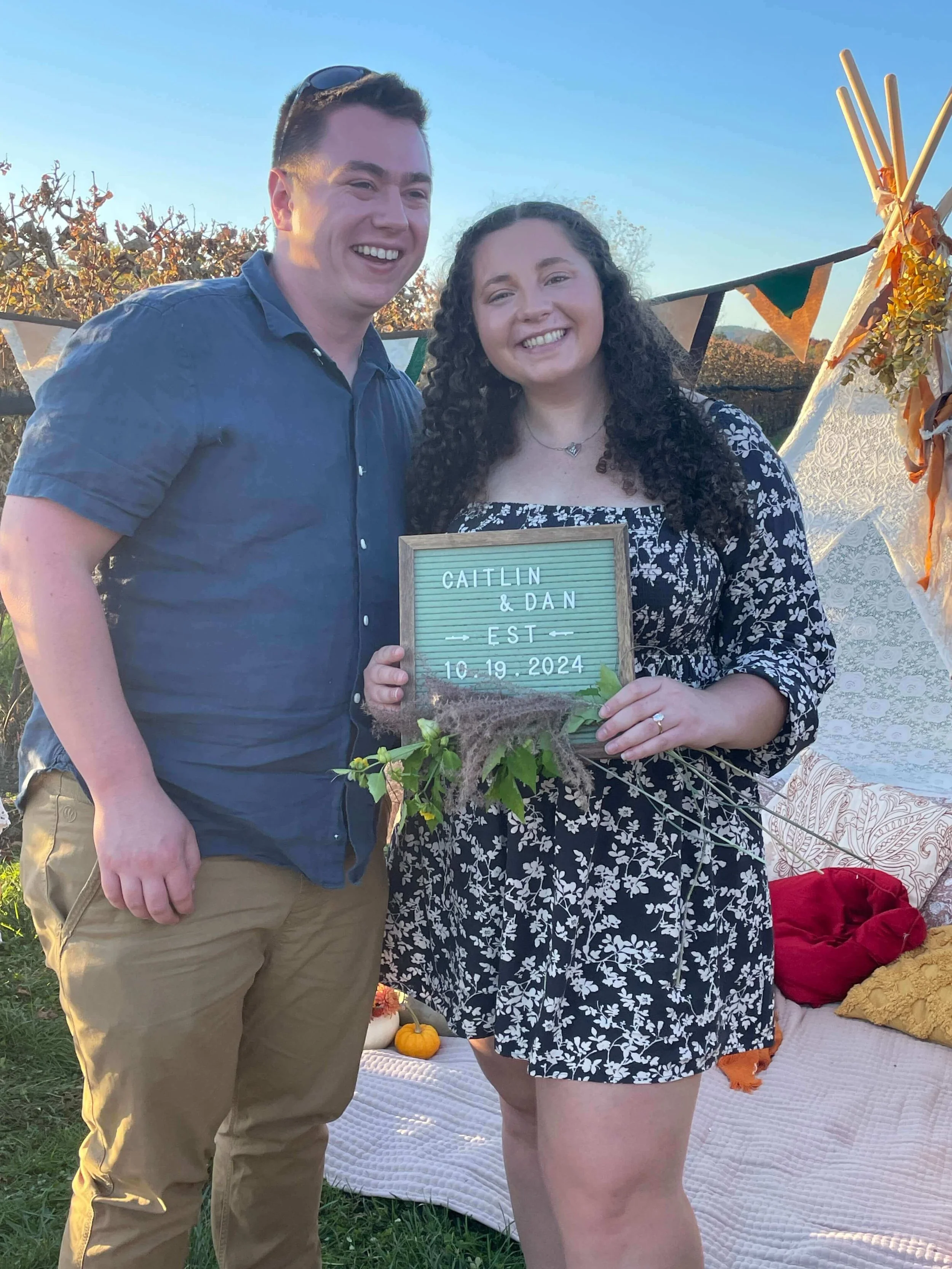 A couple celebrating outdoors at a fall event, holding a sign with their names and engagement date, Caitlin & Dan, October 19, 2024.