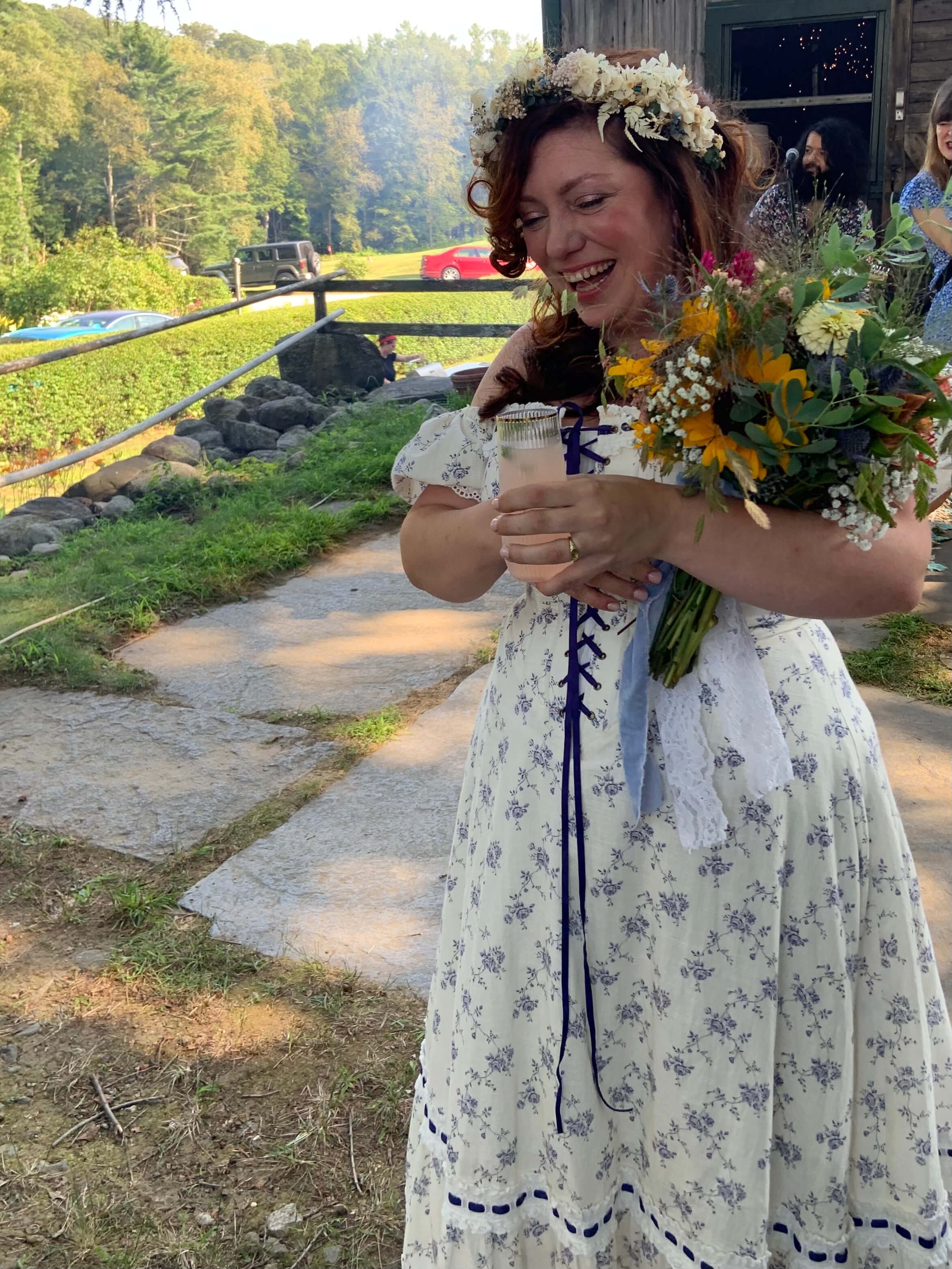 A bride wearing a white floral dress and a flower crown is smiling and holding a bouquet of flowers and a glass of champagne outdoors near a rustic building with trees in the background.