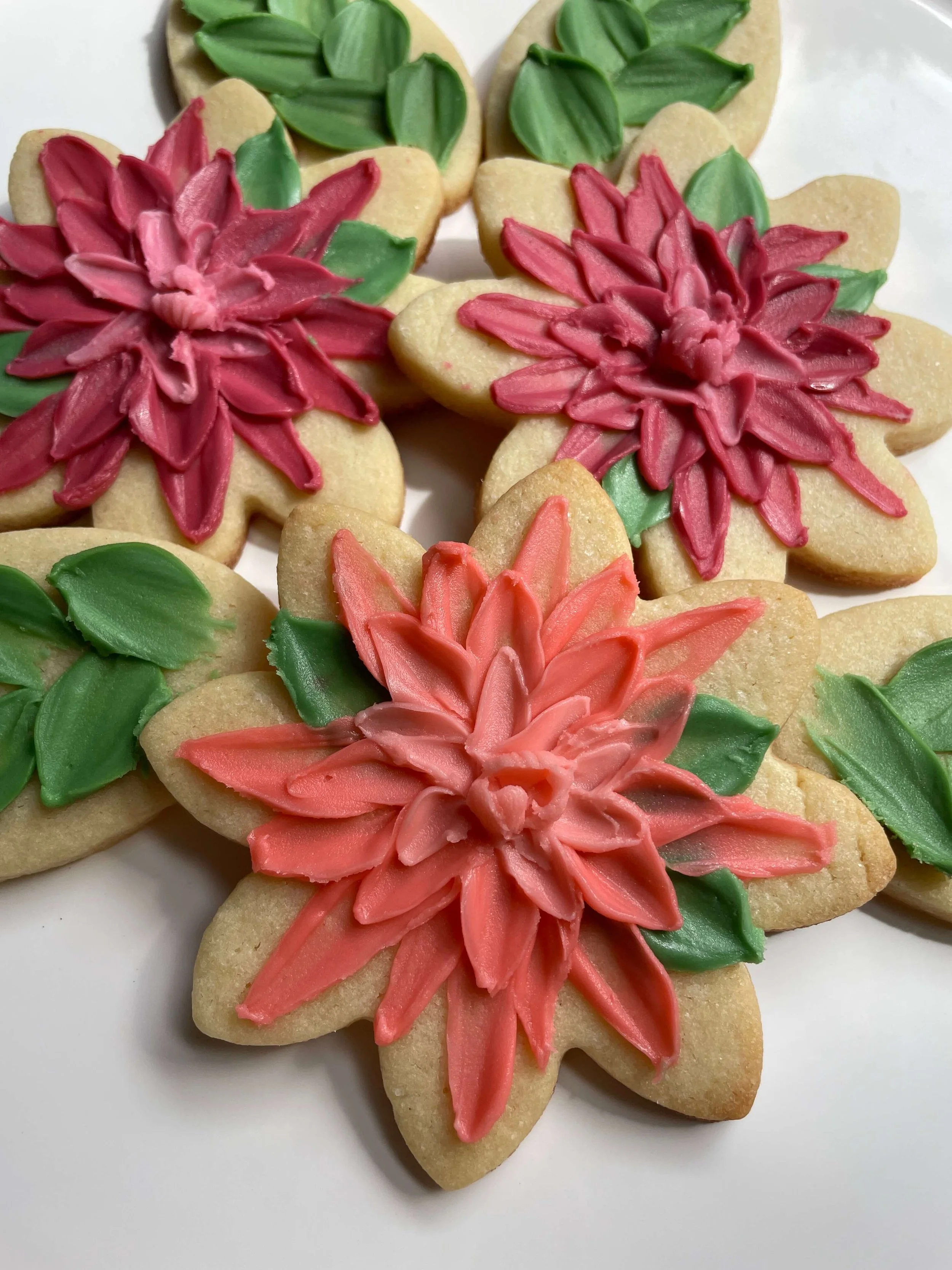 Sugar cookies decorated with pink, coral, and red icing in the shape of poinsettia flowers with green leaves.