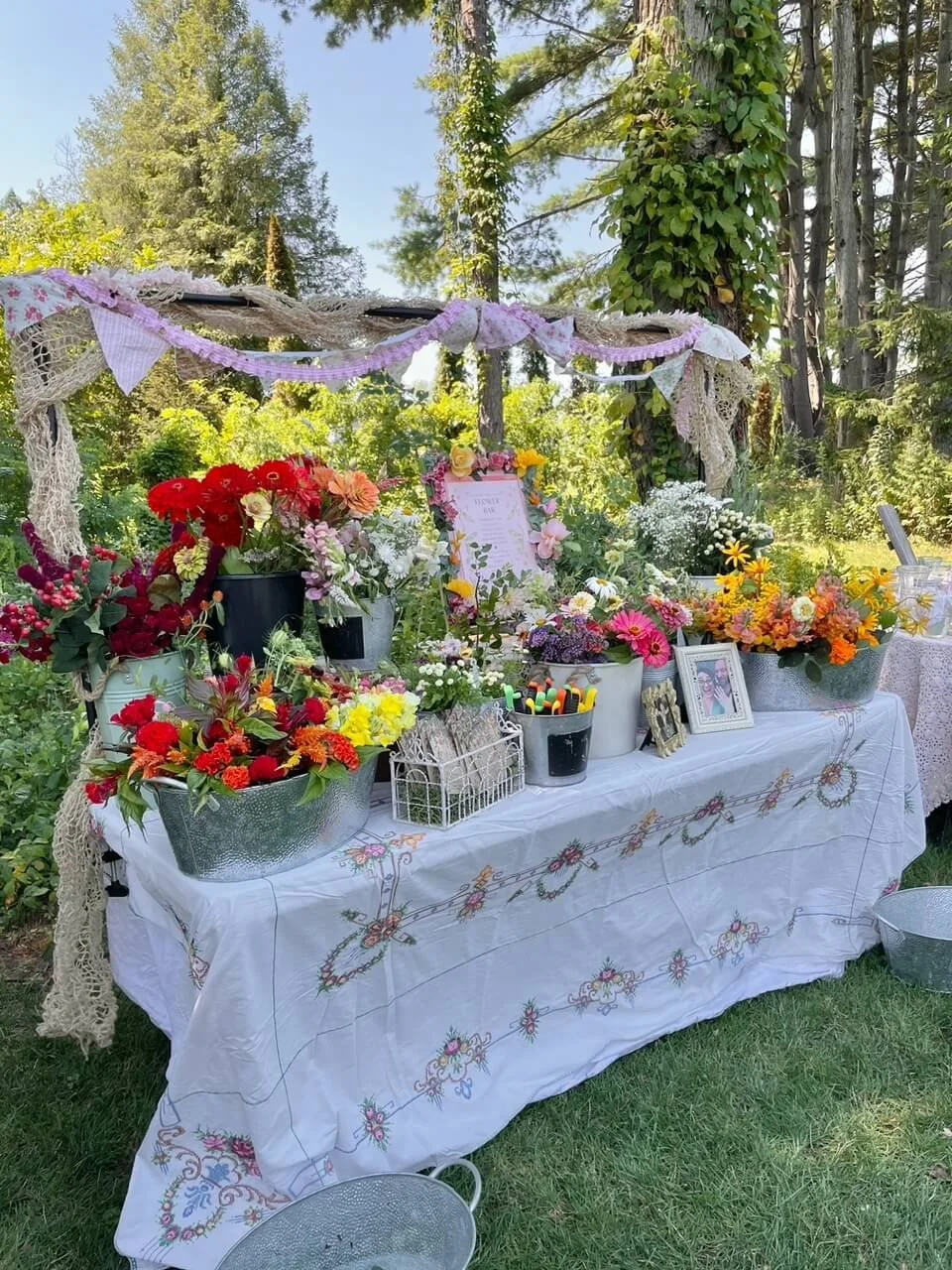 Table decorated with various colorful flowers in pots, framed photos, and small decorative items, set outdoors in a lush green garden with trees in the background.