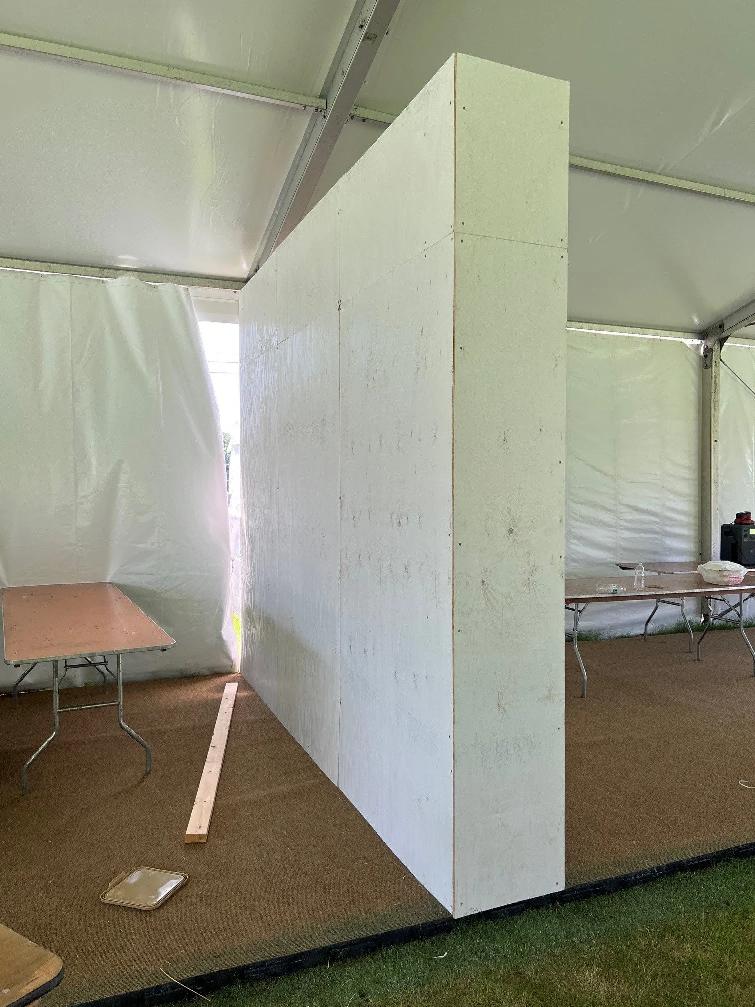 Interior of a large tent with a white wooden partition wall, a small table with materials, and a cardboard tray on a brown carpeted floor.