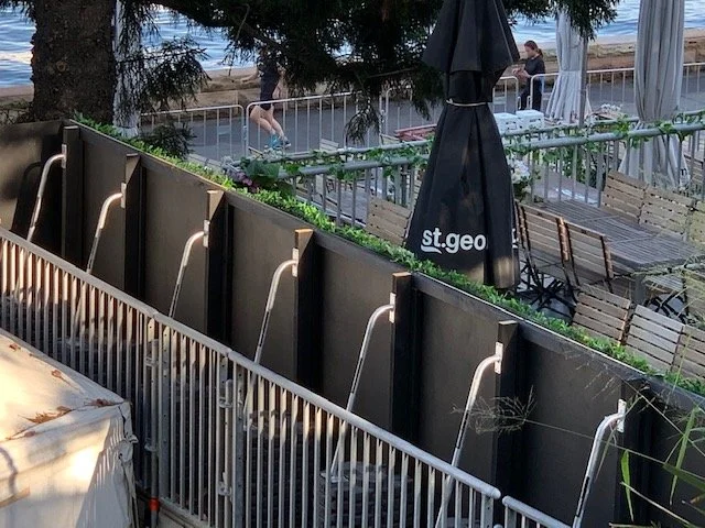 Outdoor patio area with seating, an umbrella, and a view of the beach or shoreline in the background, as well as a fenced-off section with shower stalls and metal poles.