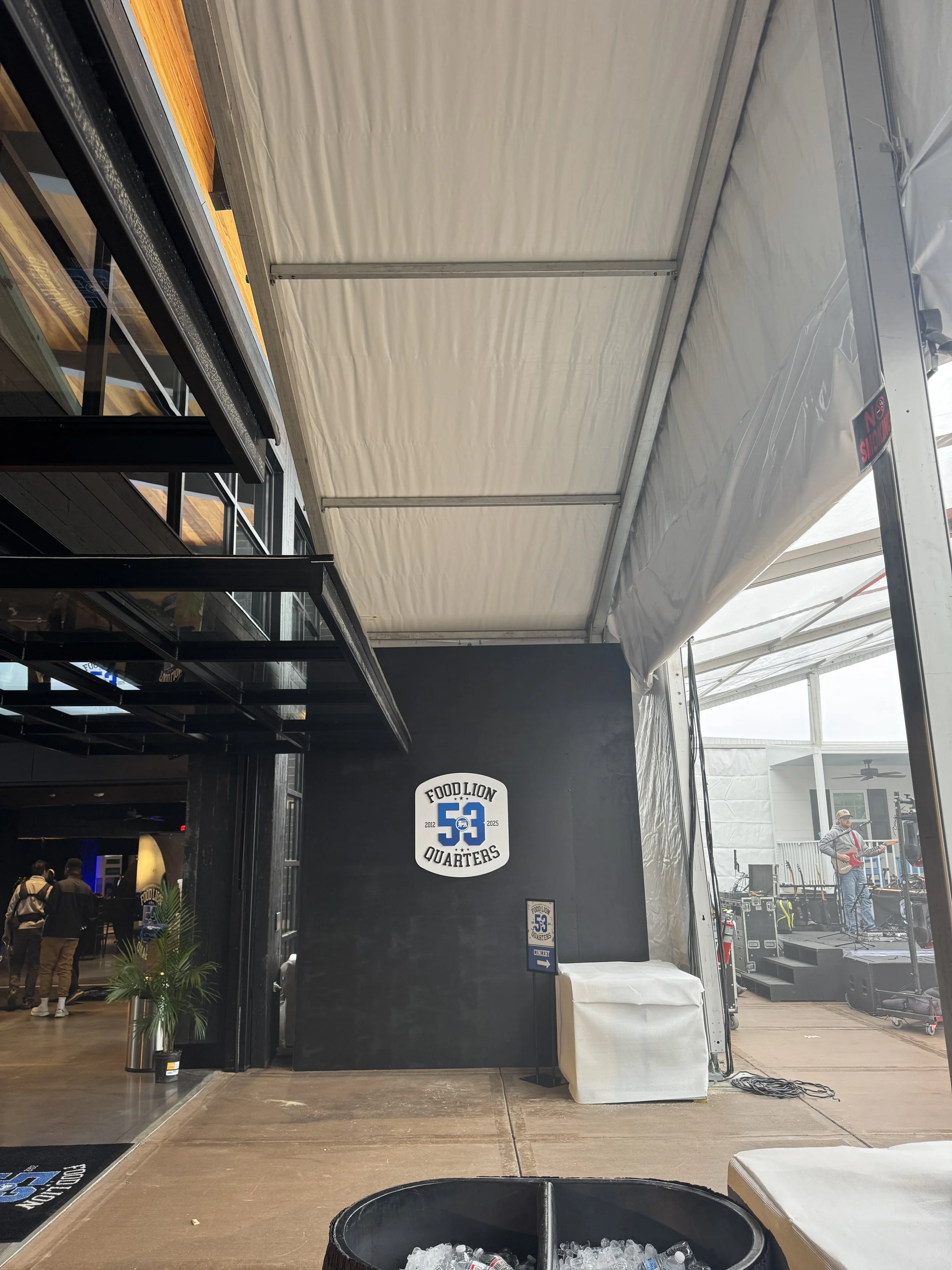 Indoor area with a stage, musical band performing, Food Lion Quarters logo on the black wall, a standee with the same logo, a trash bin filled with ice and plastic bottles, and a plant near the entrance.