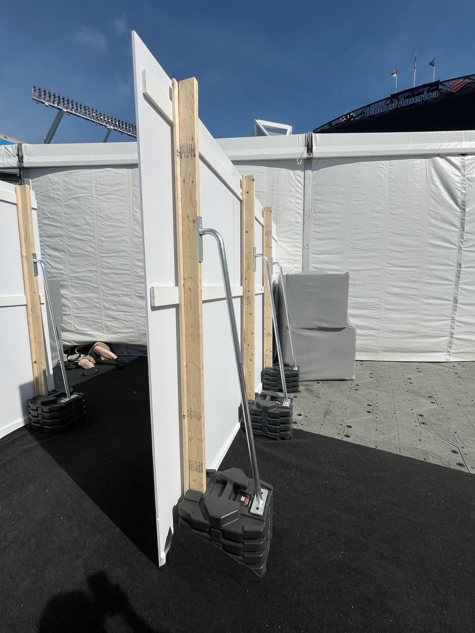 Photo of portable photo booths with white walls, wooden supports, and black rubber mats outside, under a blue sky, with part of a stadium and the Bank of America sign visible in the background.
