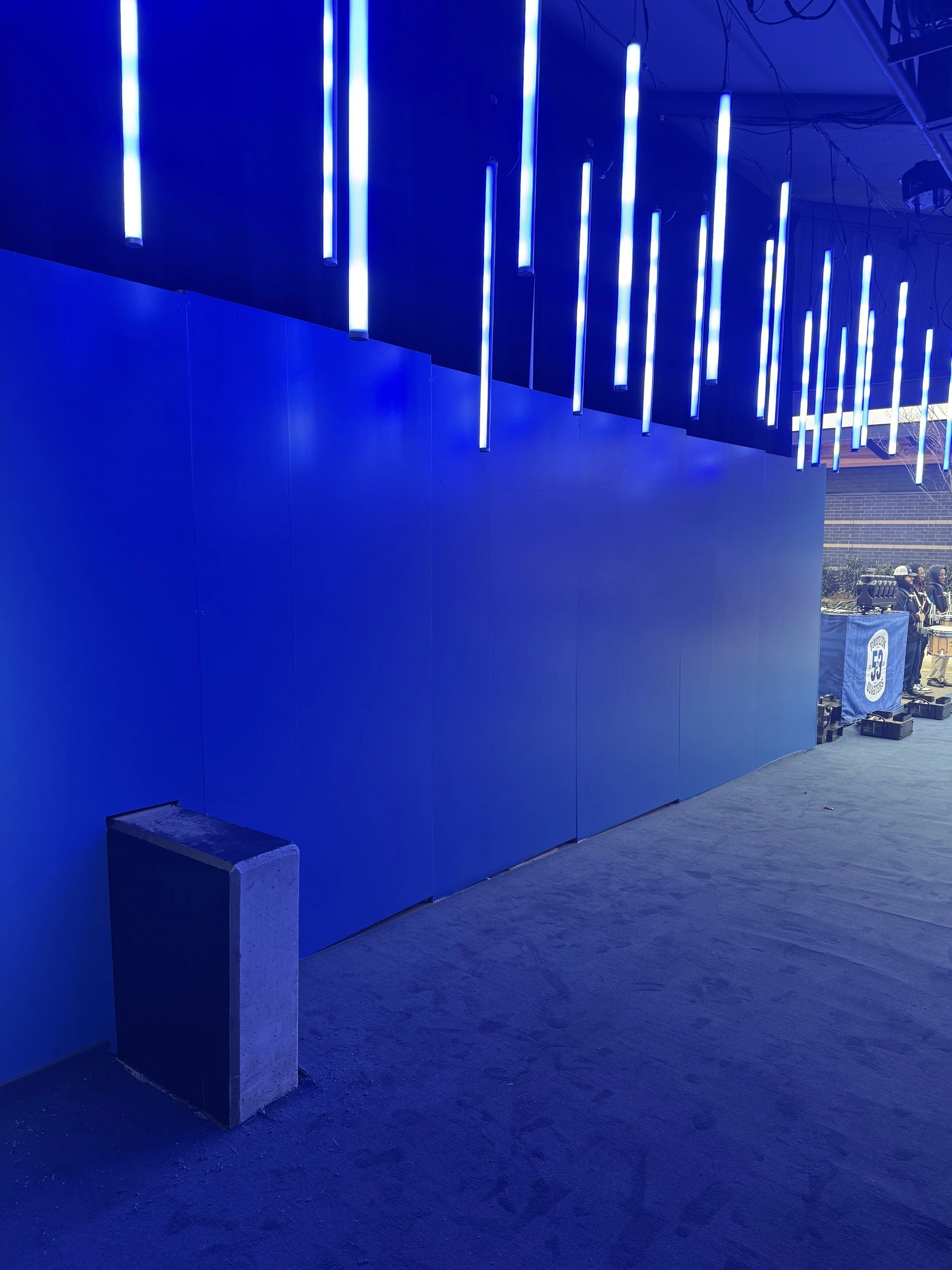An indoor space with a blue wall and ceiling decorated with vertical blue neon lights. There is a concrete block at the bottom left corner and a view of an outdoor area with people and equipment in the background.