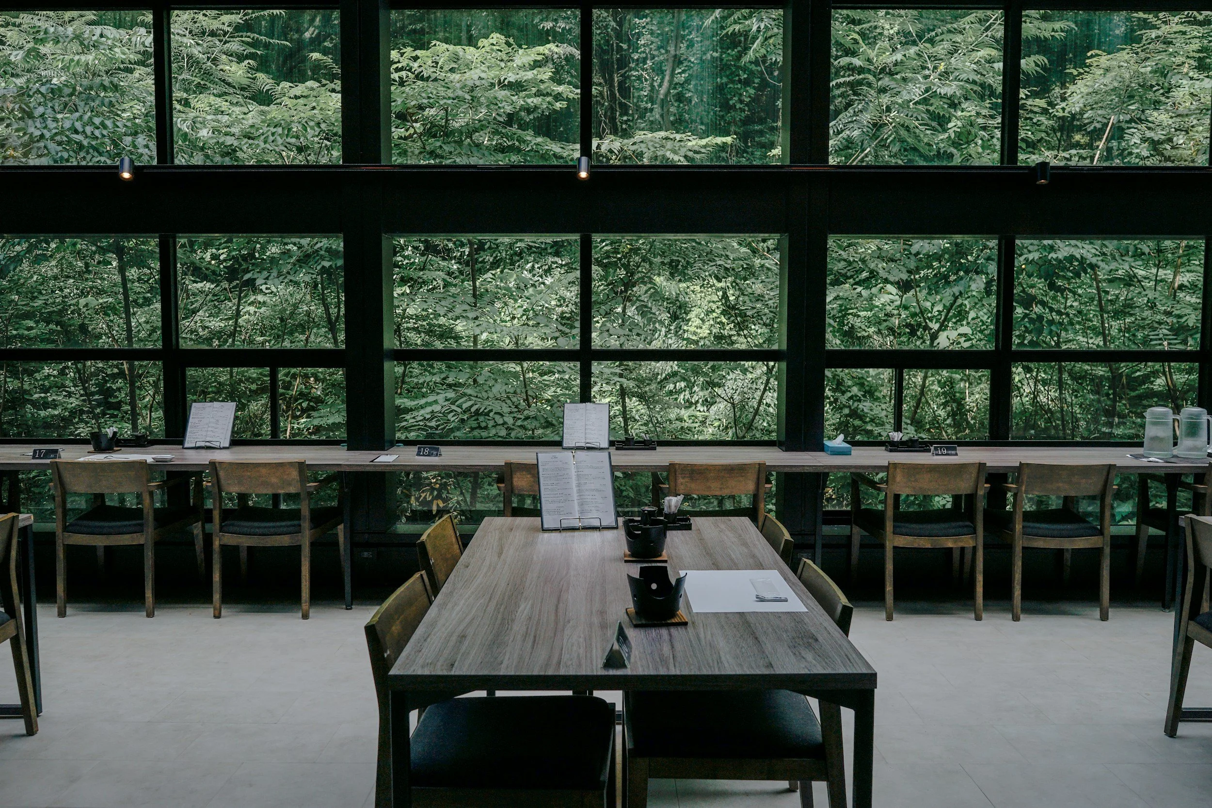 Interior of a modern restaurant or cafe with a long, narrow window offering a view of lush greenery outside. There is a wooden table with chairs around it, some menus, and water pitchers on a counter along the window.