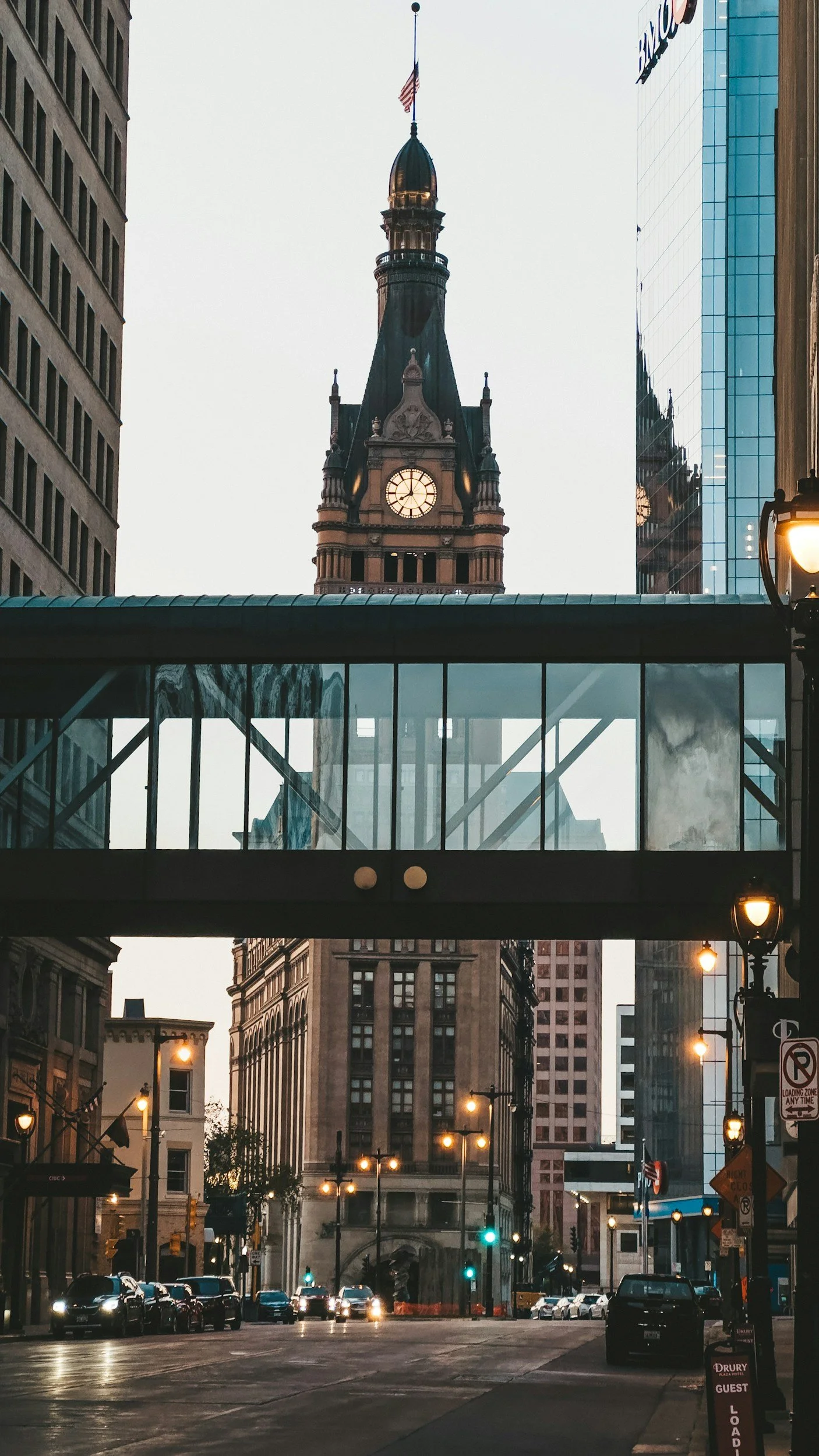 A city street scene with tall buildings, street lamps, parked cars, and a prominent historic clock tower with a green roof and an American flag on top, visible behind a glass pedestrian bridge.