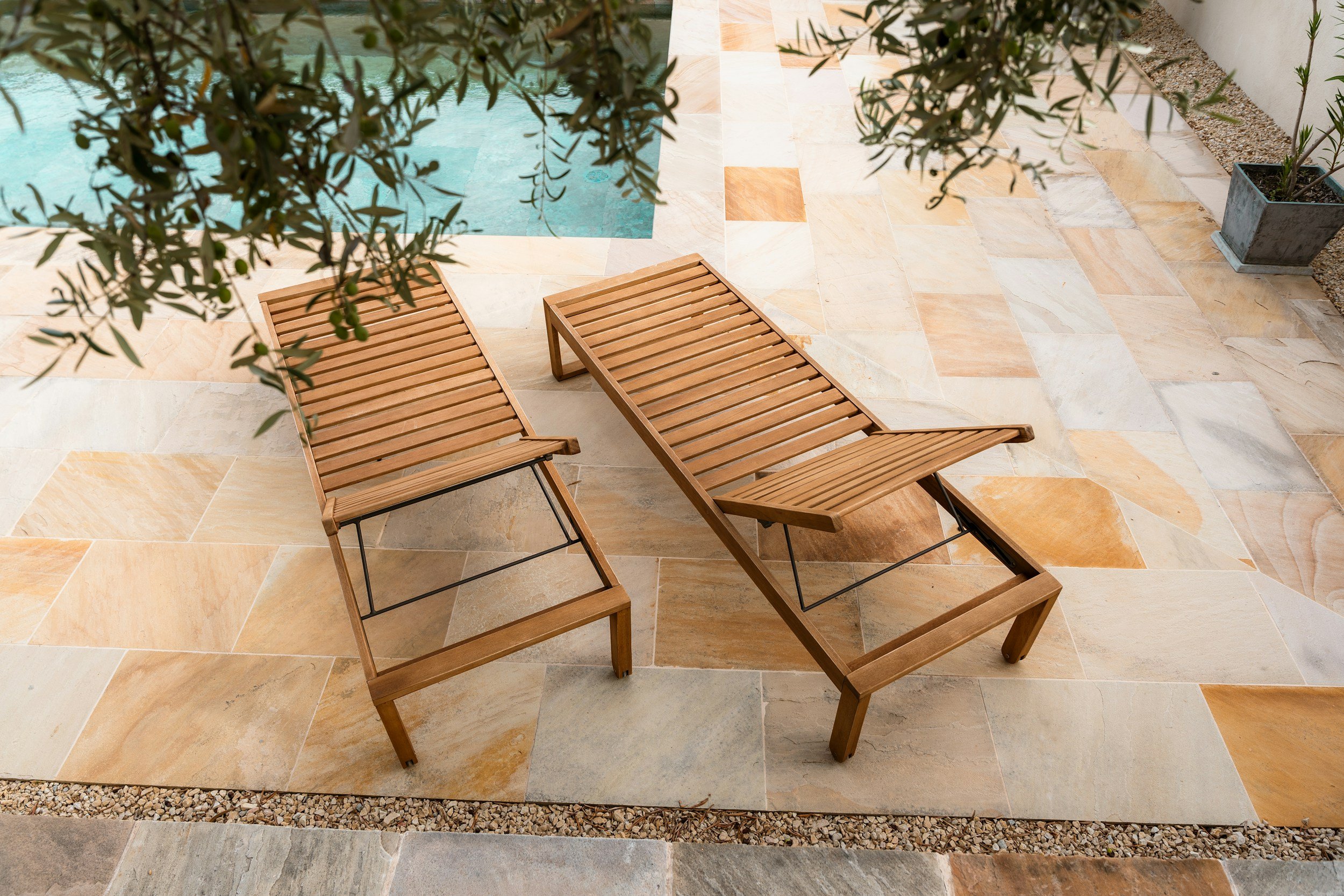 Two wooden lounge chairs by a swimming pool on a tiled patio with potted plants nearby.