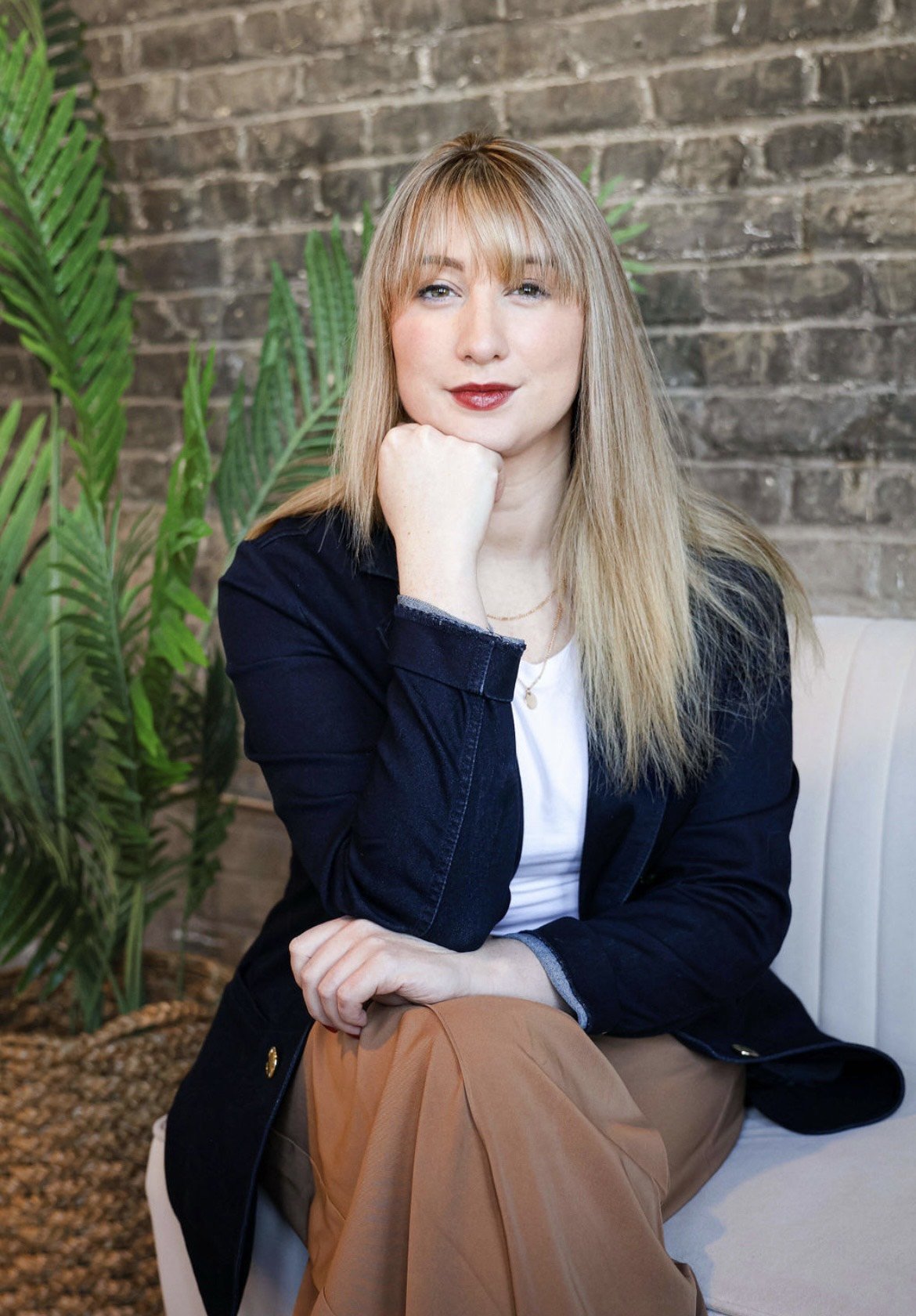 A woman with blonde hair and bangs, wearing a black blazer and white shirt, sitting on a white chair with a brick wall behind her and green plants on the side.