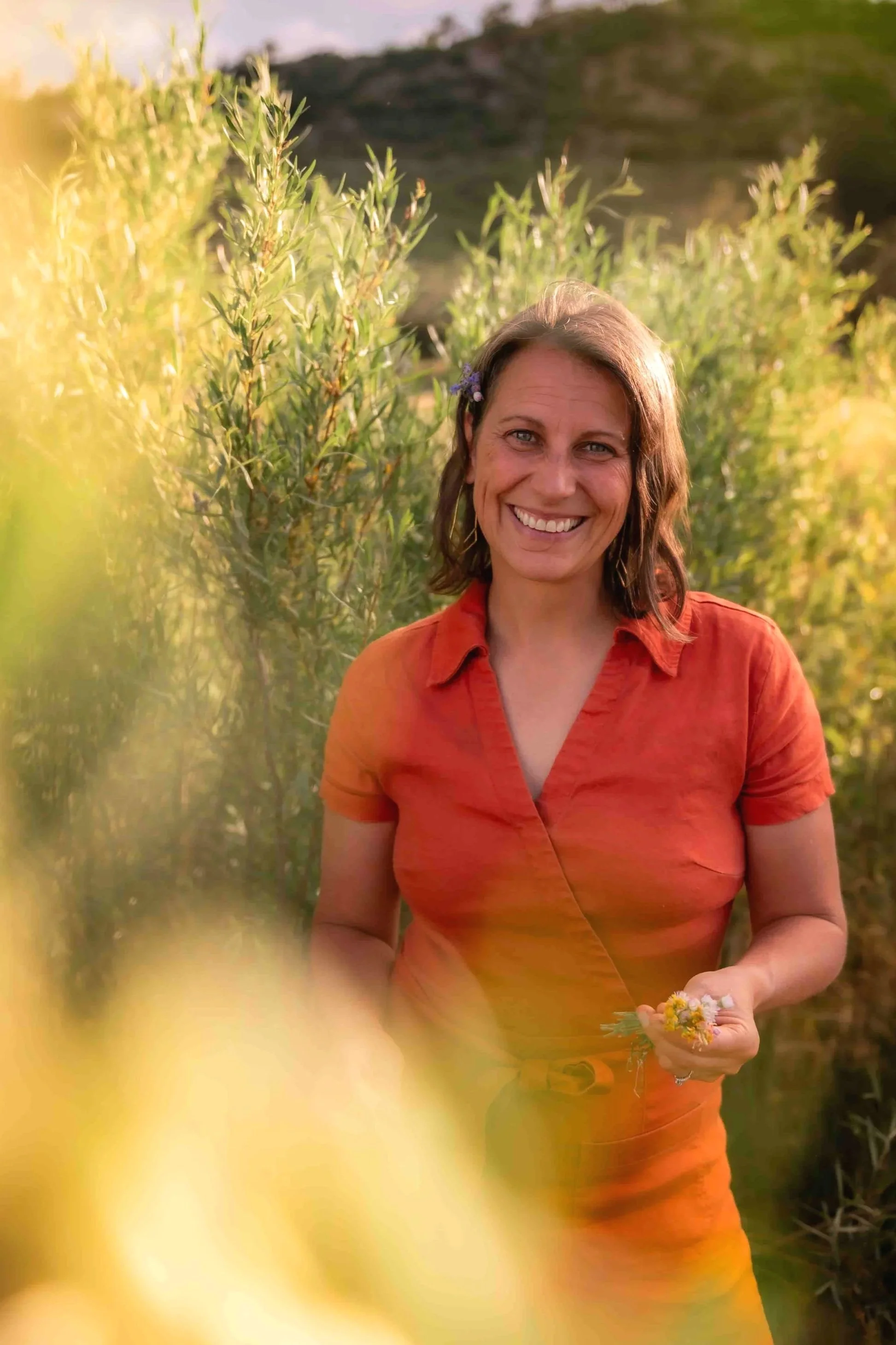 Photo of Heather in a cute orange jumpsuit, smiling at the camera holding wildflowers and with bush leaves in the foreground