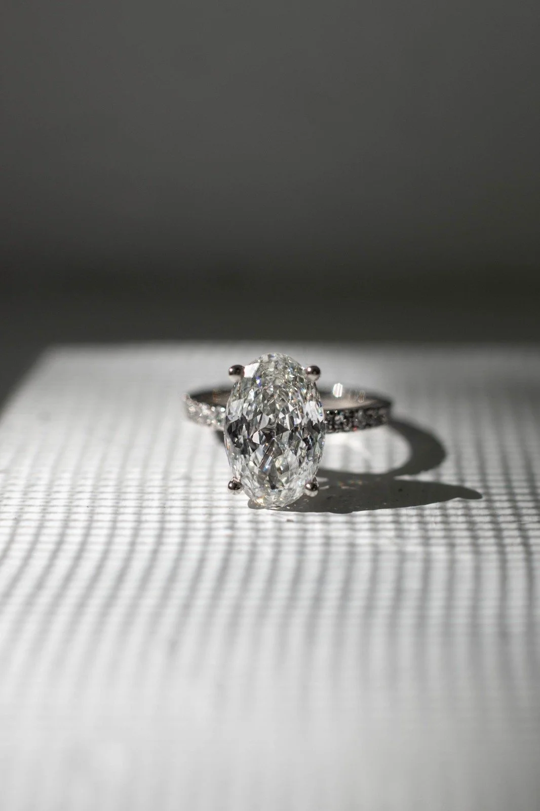 A close-up of a silver ring with a large, oval-cut diamond centered on a textured white surface. The diamond casts a shadow, with a plain gray background.