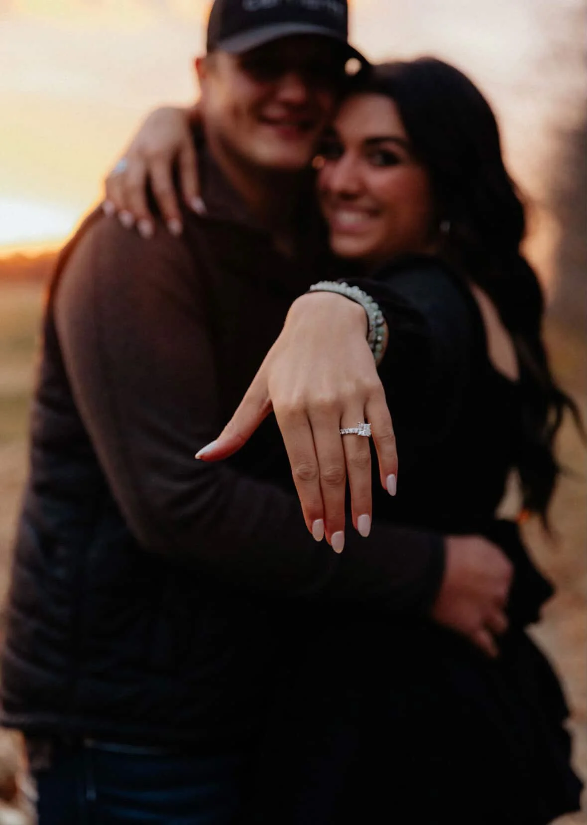 A woman showing her engagement ring with a sunset in the background, embracing a man.