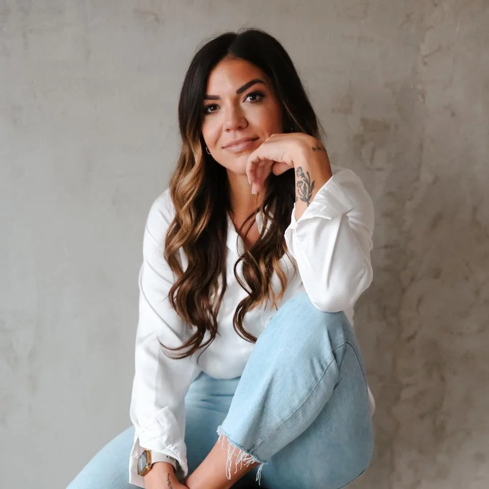 A woman with long brown hair and tattoos on her hand is sitting against a neutral wall, resting her chin on her hand and looking at the camera.