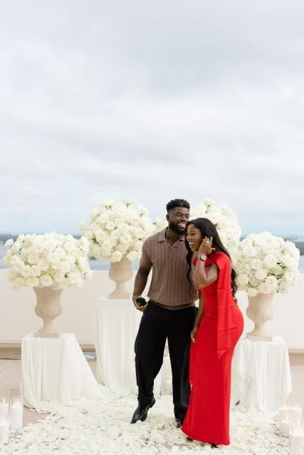 A smiling man and woman standing outdoors in front of large white floral arrangements, with the man holding a smartphone, on a cloudy day.