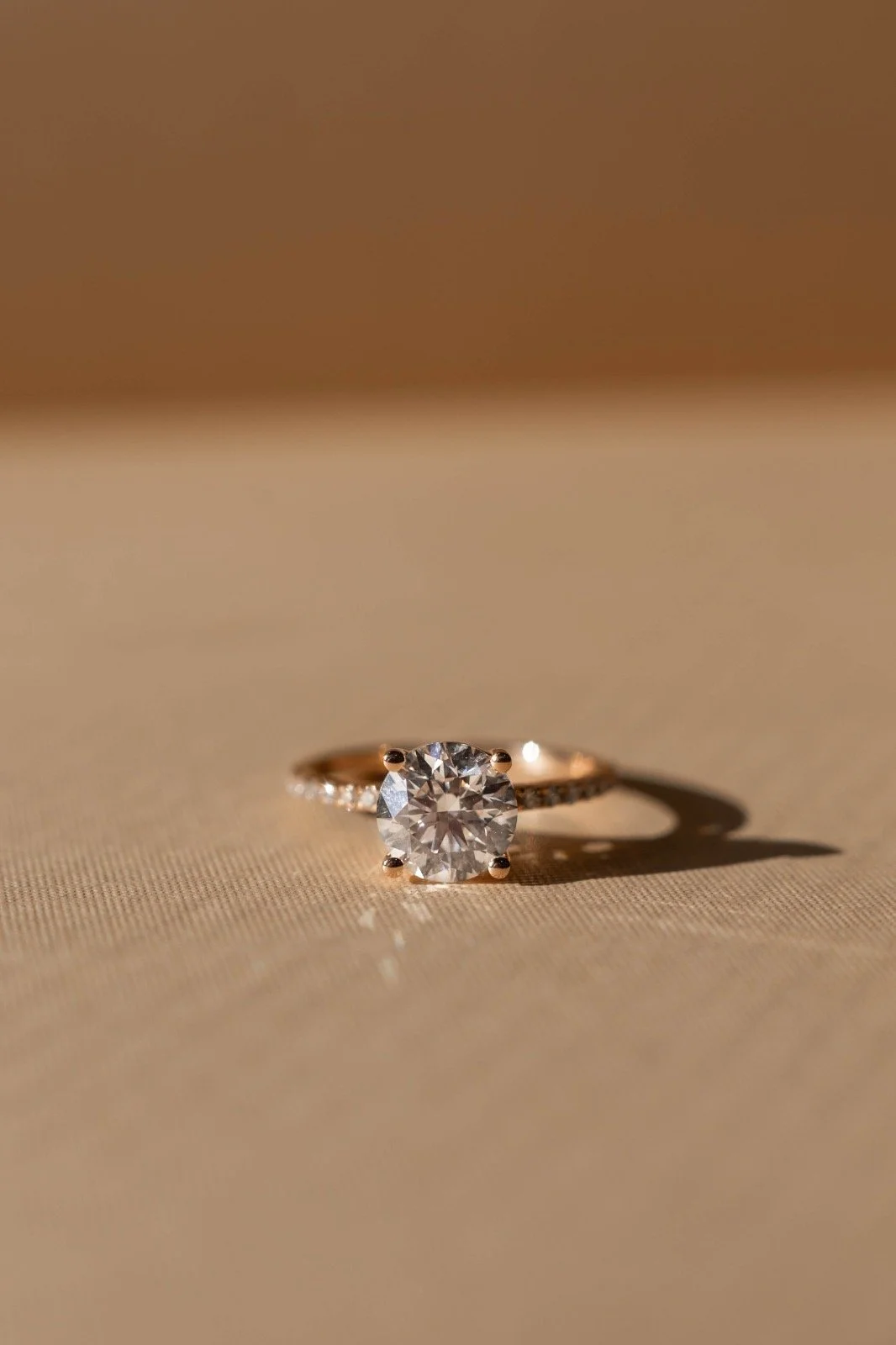 Close-up of an engagement ring with a large central diamond on a beige surface, casting a shadow.