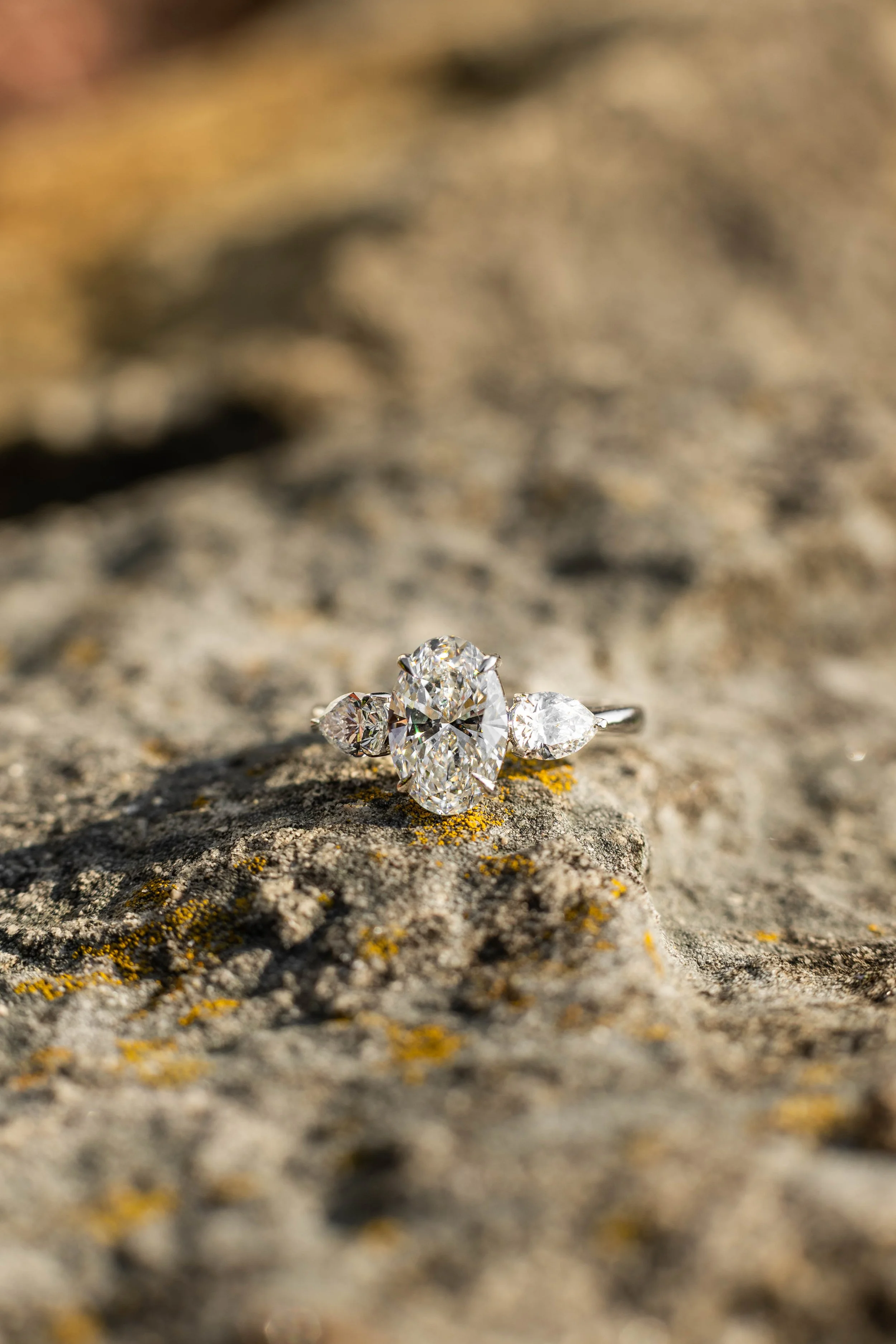 Close-up of a diamond ring with a large oval diamond and smaller side diamonds, placed on a textured rock surface outdoors.