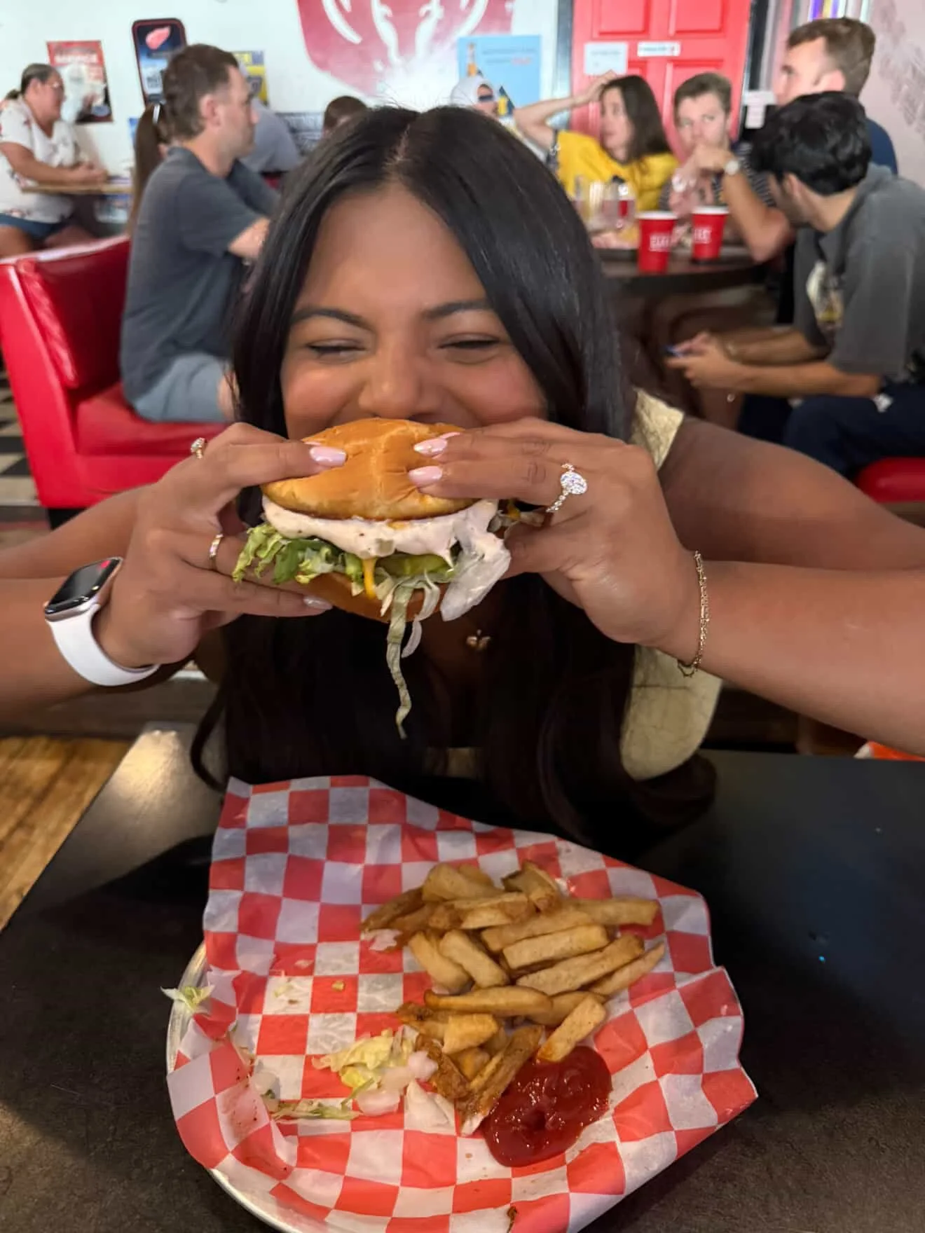 Woman with dark hair and jewelry eating a burger with lettuce and a bun at a restaurant. On the table, there is a basket of French fries with ketchup. The background shows a lively restaurant with several people seated and talking.