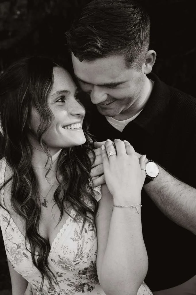 A couple sharing an intimate moment: woman smiling and looking into man's eyes, hands clasped with rings visible, close-up black and white photograph.
